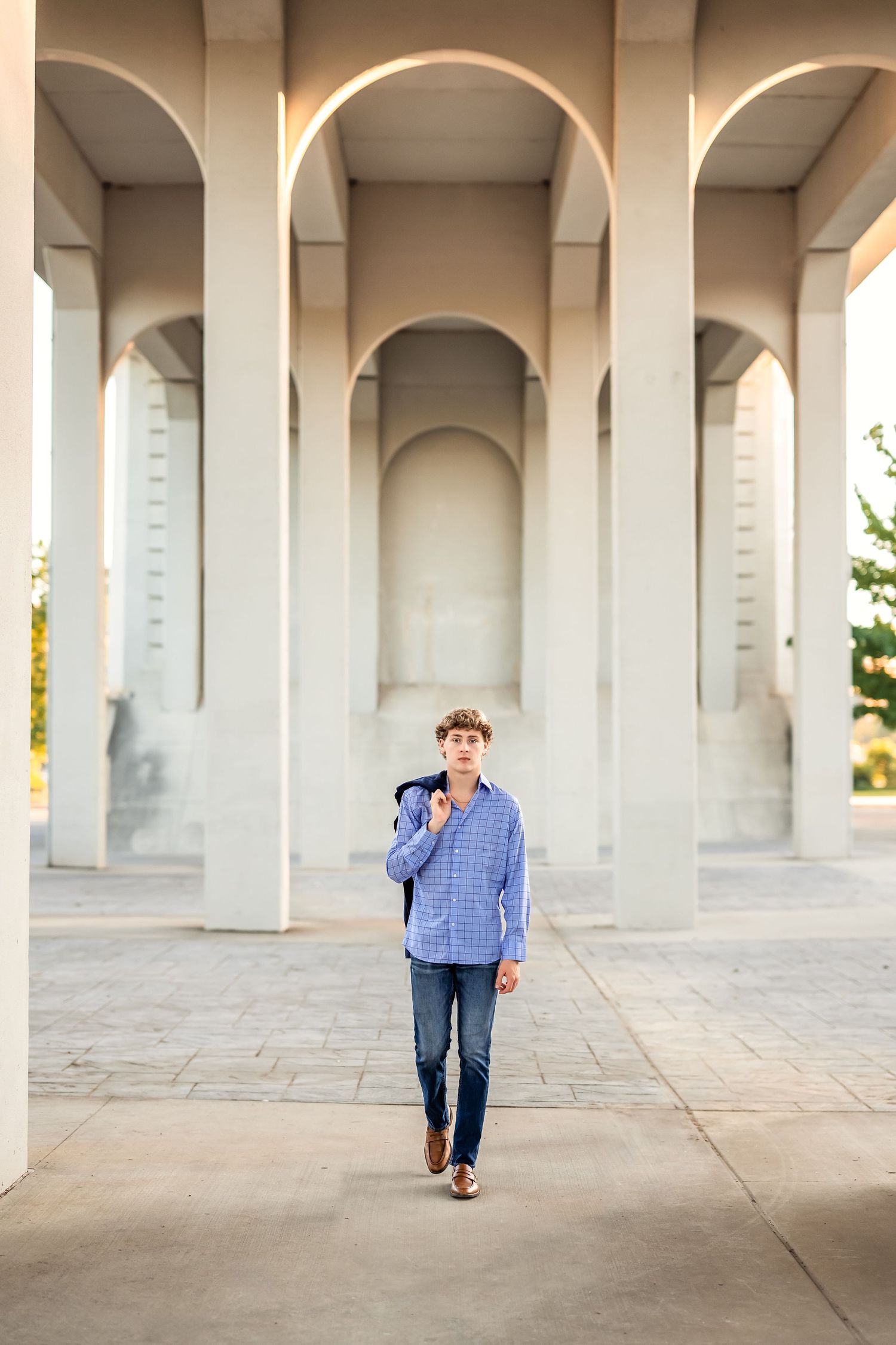 Senior boy holds jacket over shoulder and walks under a set of white concrete arches in a repeating architectural pattern in evening light