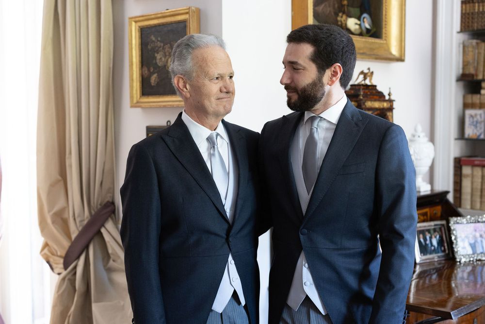 Two men in dark suits stand in an elegant room with tan curtains and framed artwork on the wall.
