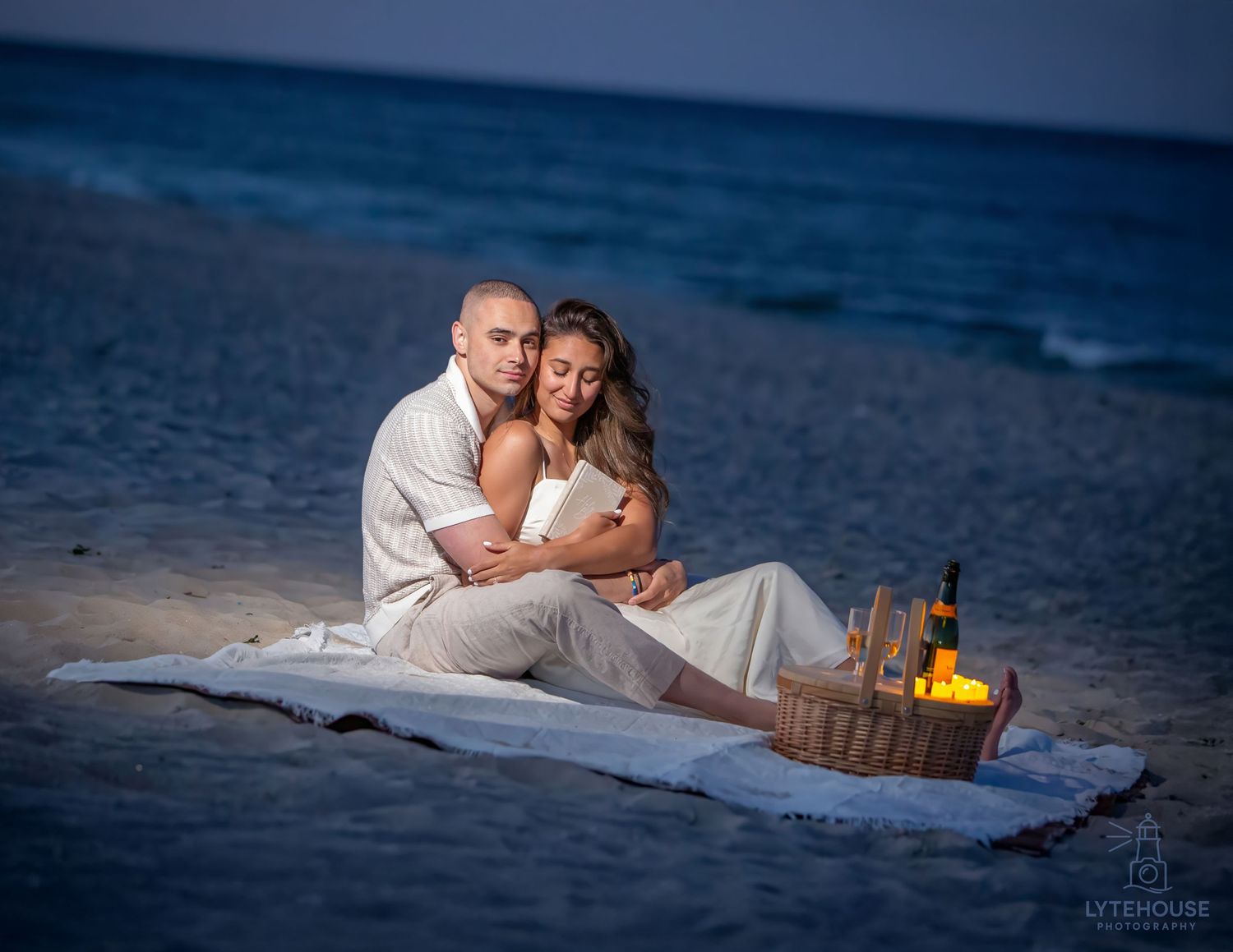 Couple in casual beach attire poses together at dusk on the sand with houses visible in the background.
