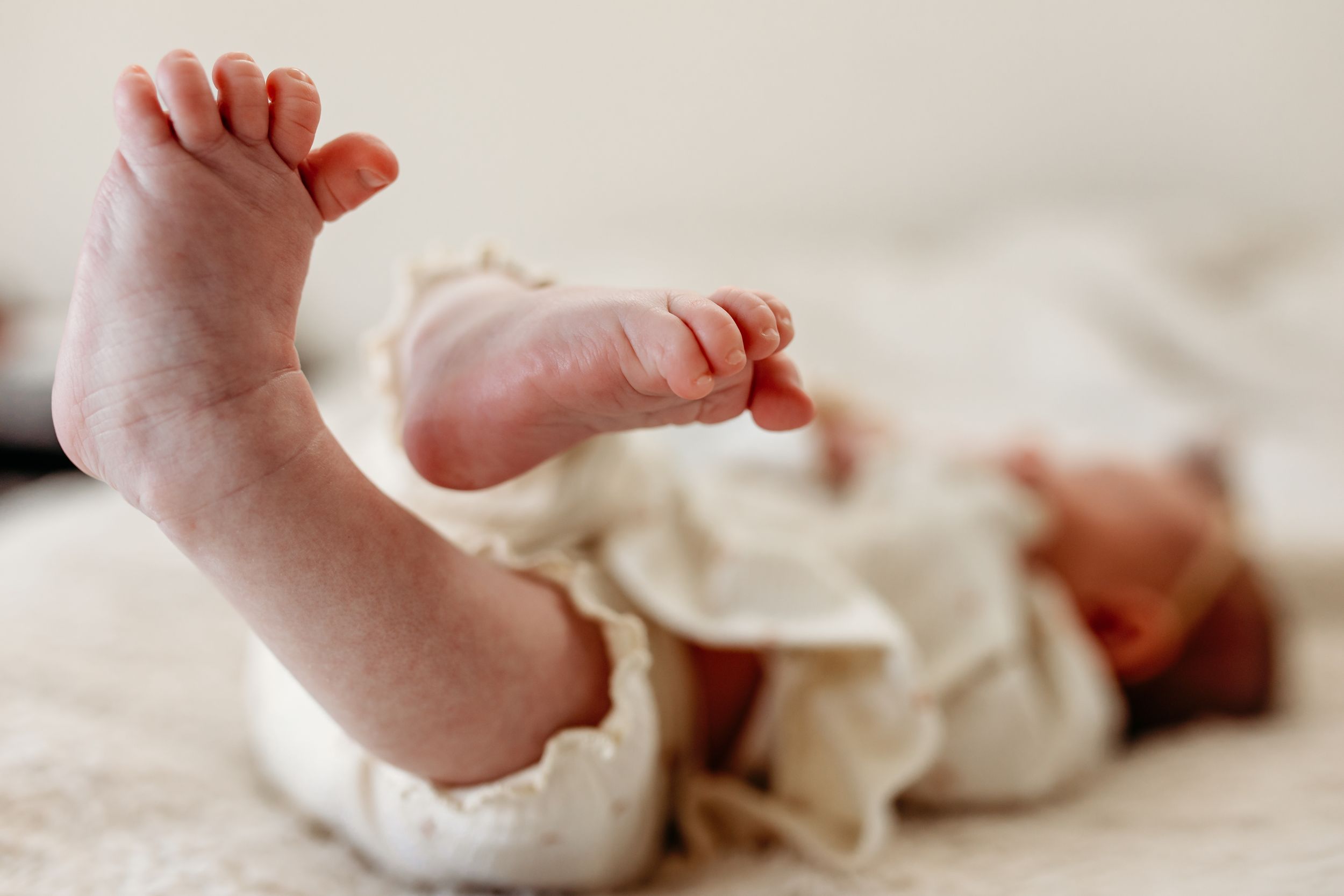 Tiny newborn baby feet with delicate toes resting on cream-colored fabric in natural light.