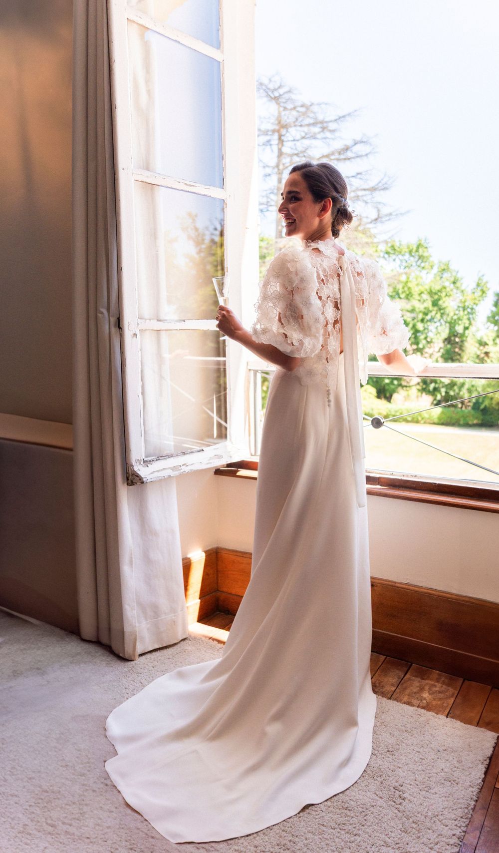 Elegant wedding dress with delicate lace sleeves and flowing train shown near a sunlit window.