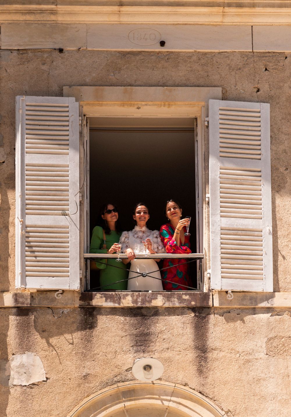 People lean out of four white-shuttered windows in an old stone building wall, creating a playful panoramic scene.