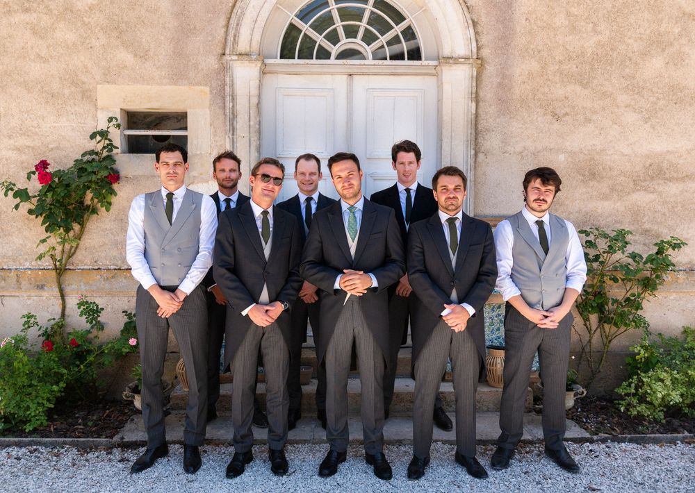 Group of wedding party members in formal attire sitting on a stone bench against a rustic wall.