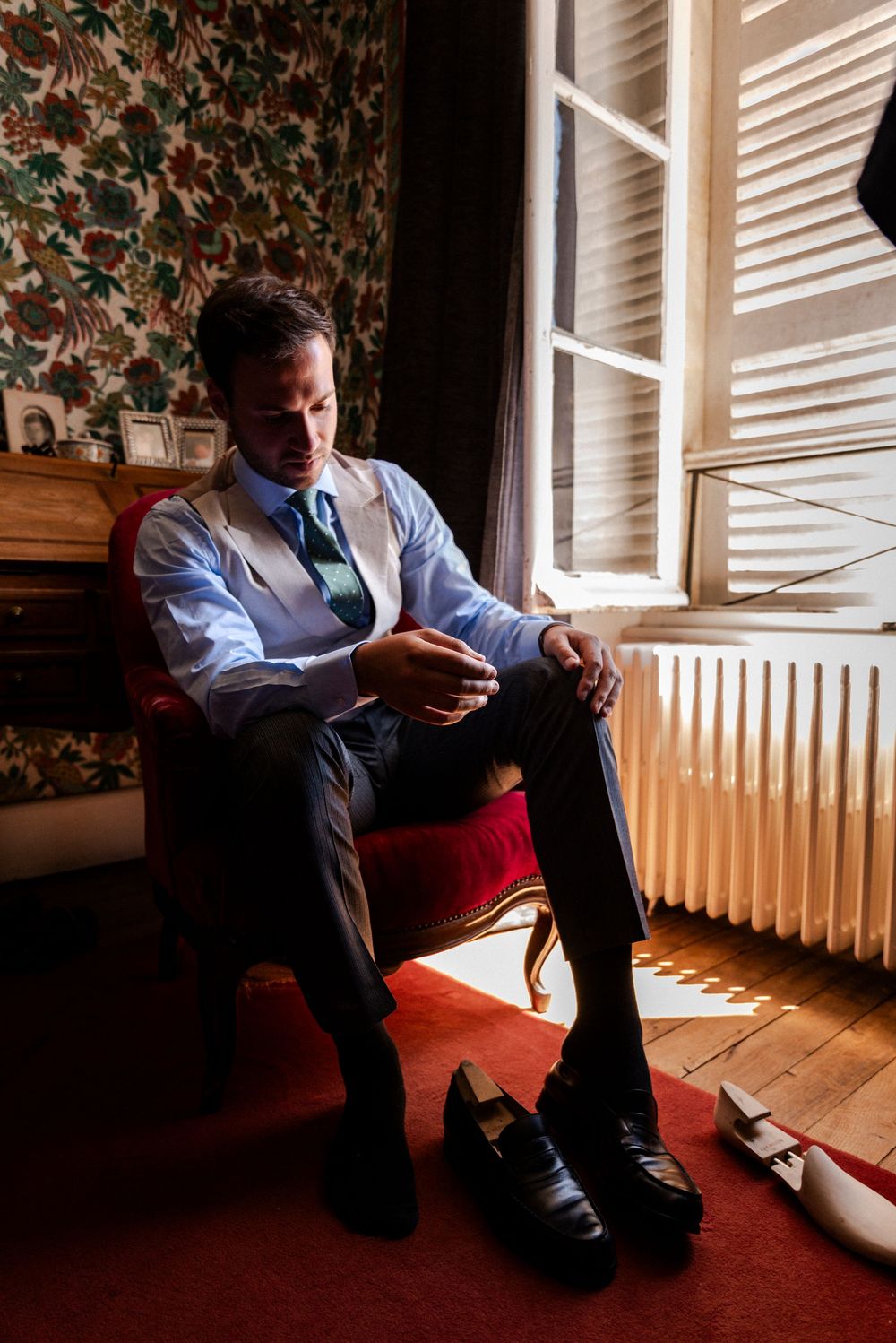 Person getting dressed in formal attire while sitting in chair by window with dress shoes nearby.