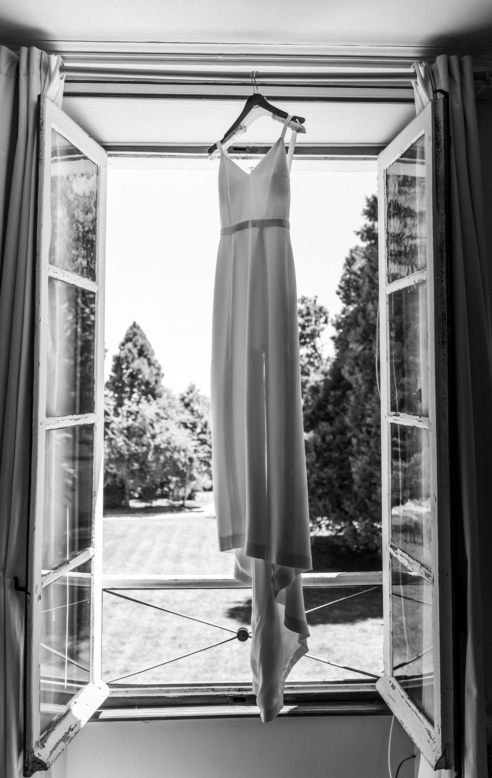 A white wedding dress hangs in an open window frame overlooking a garden in black and white photography.