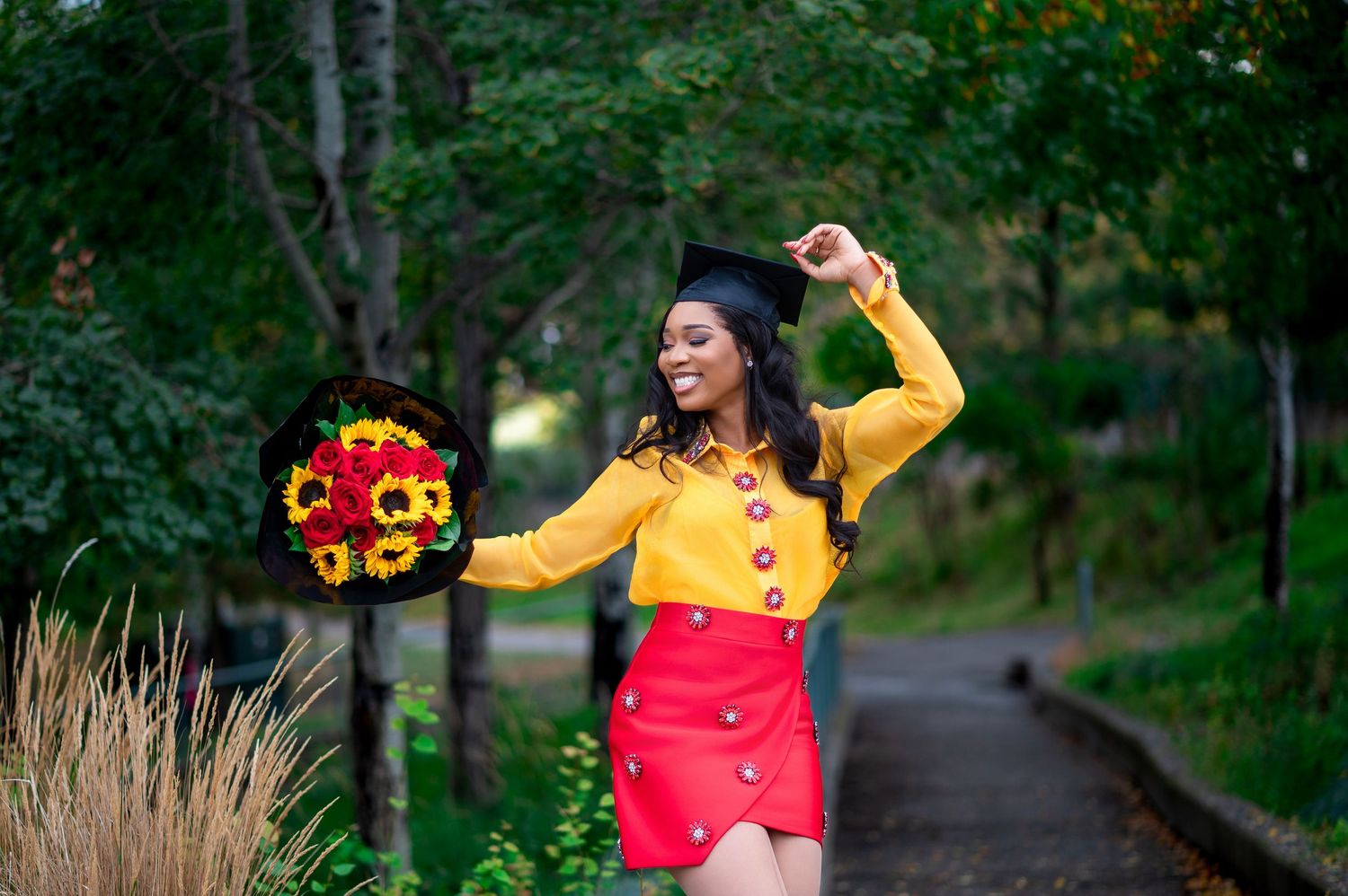 Graduate in bright yellow top and red skirt holds colorful sunflower bouquet during outdoor photoshoot.
