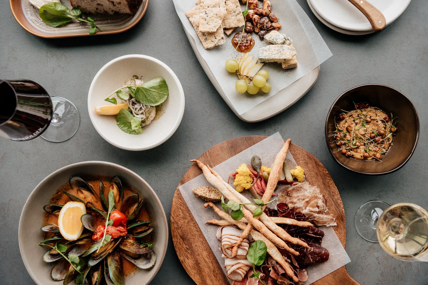 Overhead view of elegant dinner spread featuring seafood, grilled meat, charcuterie board and wine glasses on wooden table by August & Vine