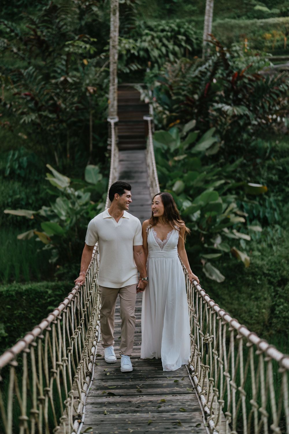 Couple walks hand in hand across rustic wooden bridge in tropical setting.