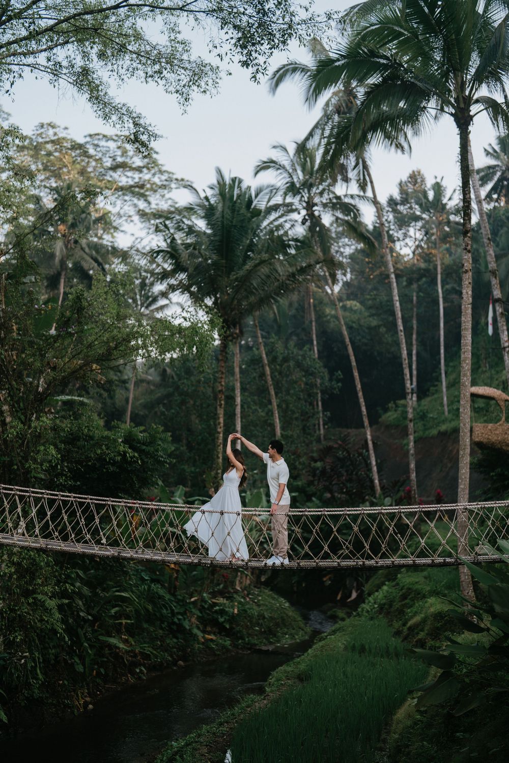 Couple in white dresses poses on suspension bridge surrounded by tropical forest.