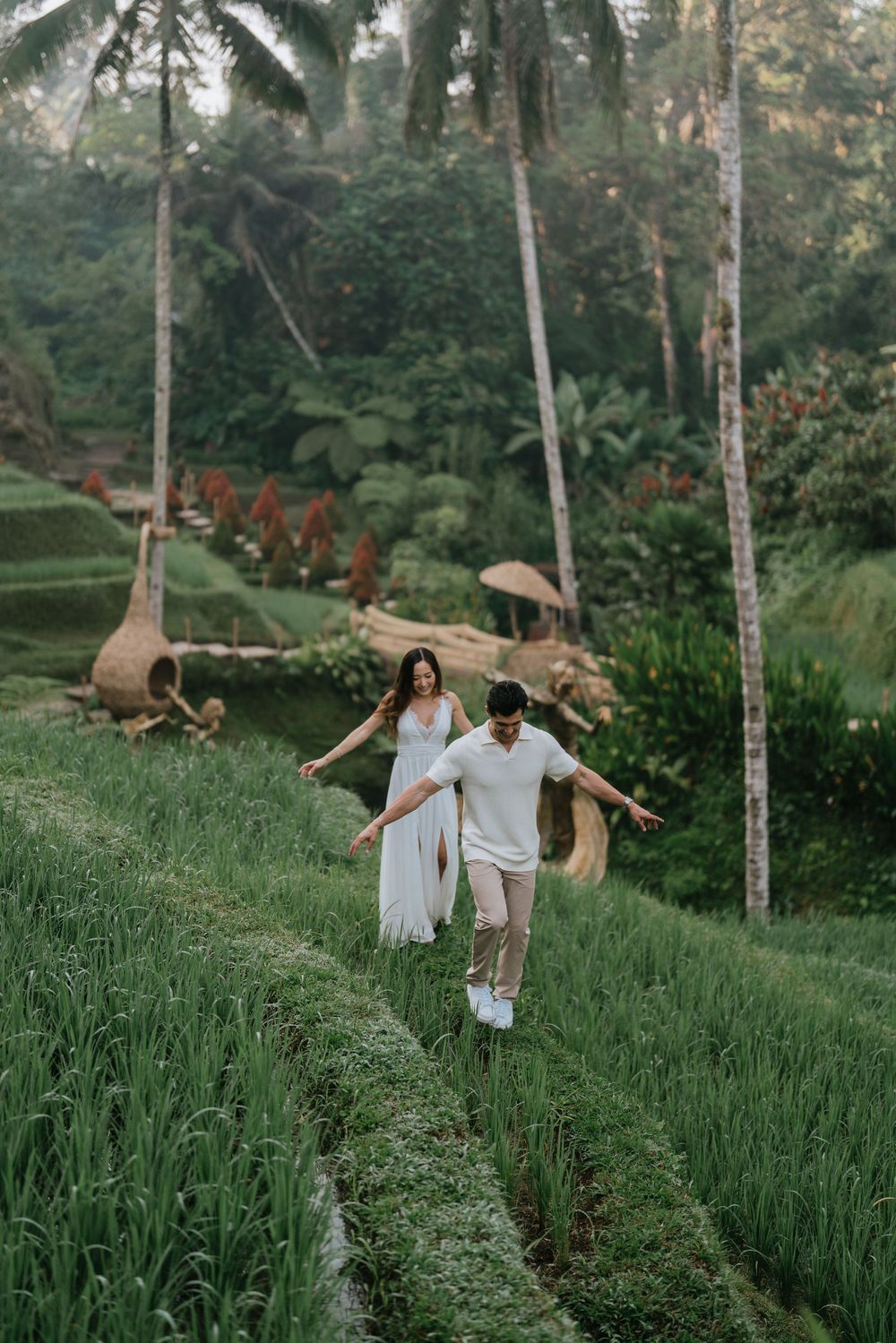 Couple in white clothing walks through tall grass field with palm trees.