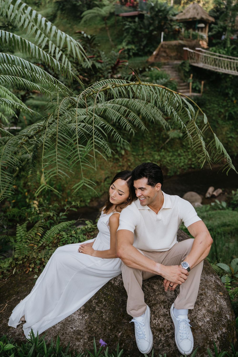 Couple in white relaxes on large rock surrounded by tropical ferns.
