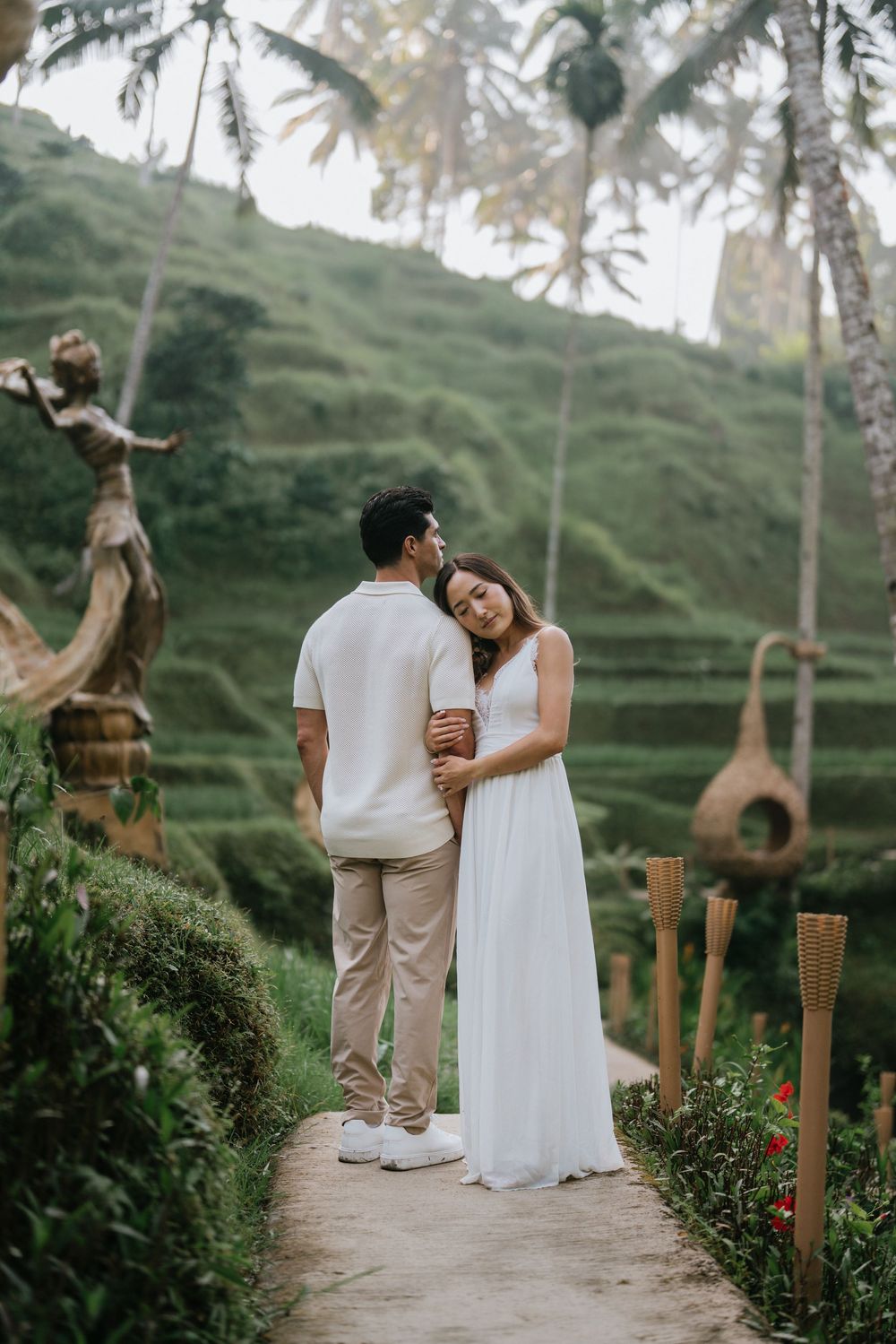 A couple in white outfits standing on a path surrounded by Balinese rice terraces and palm trees.
