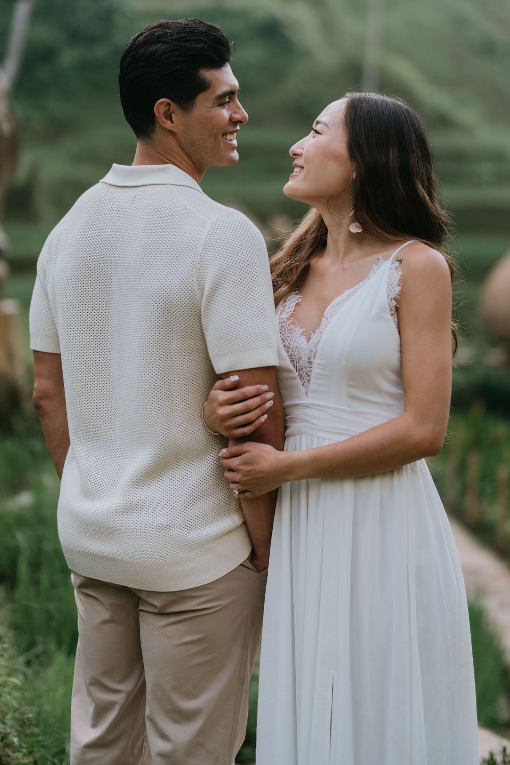 A couple in coordinated white and beige outfits share a tender moment on a tropical path.