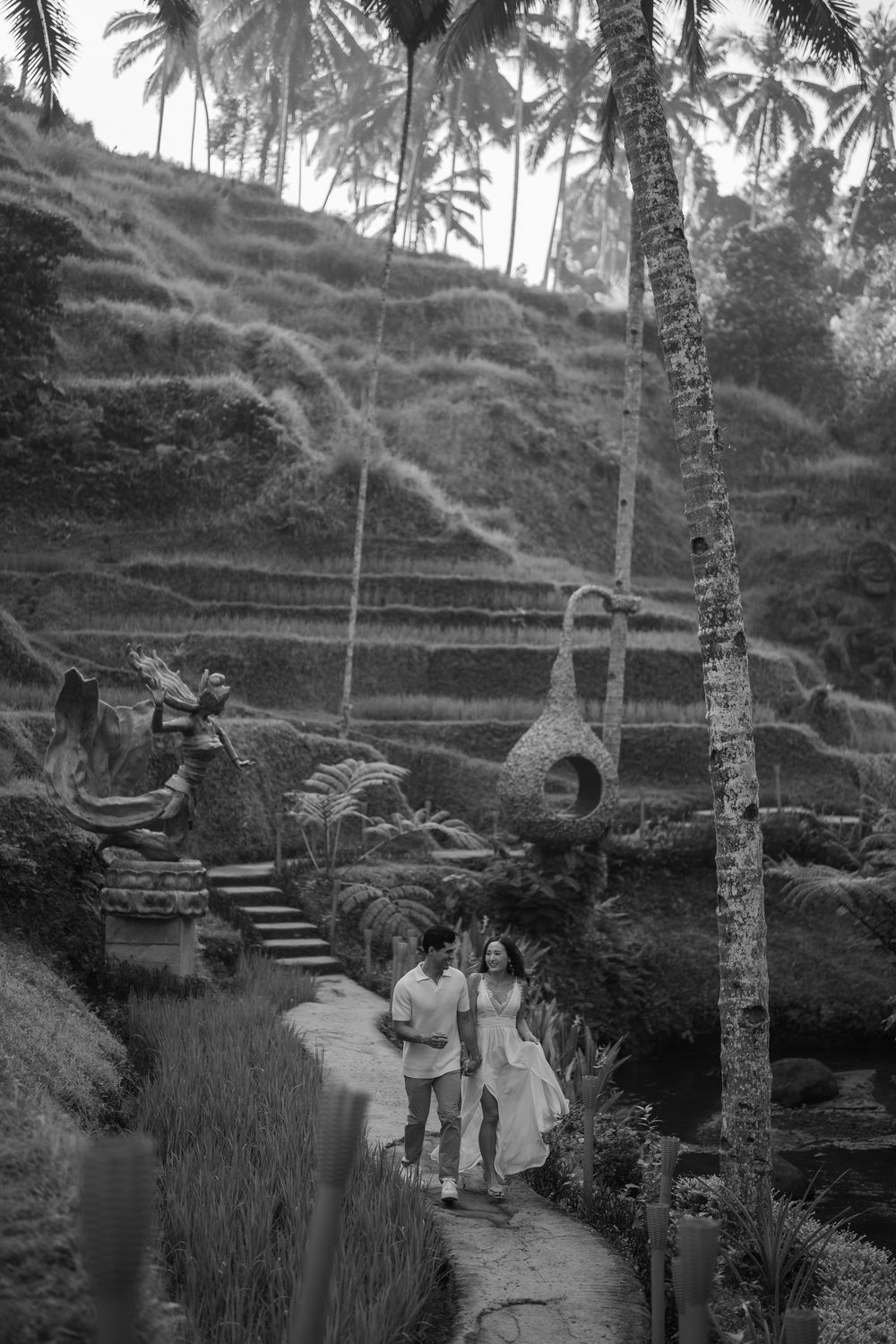A black and white photo of a couple walking along terraced rice fields with palm trees.