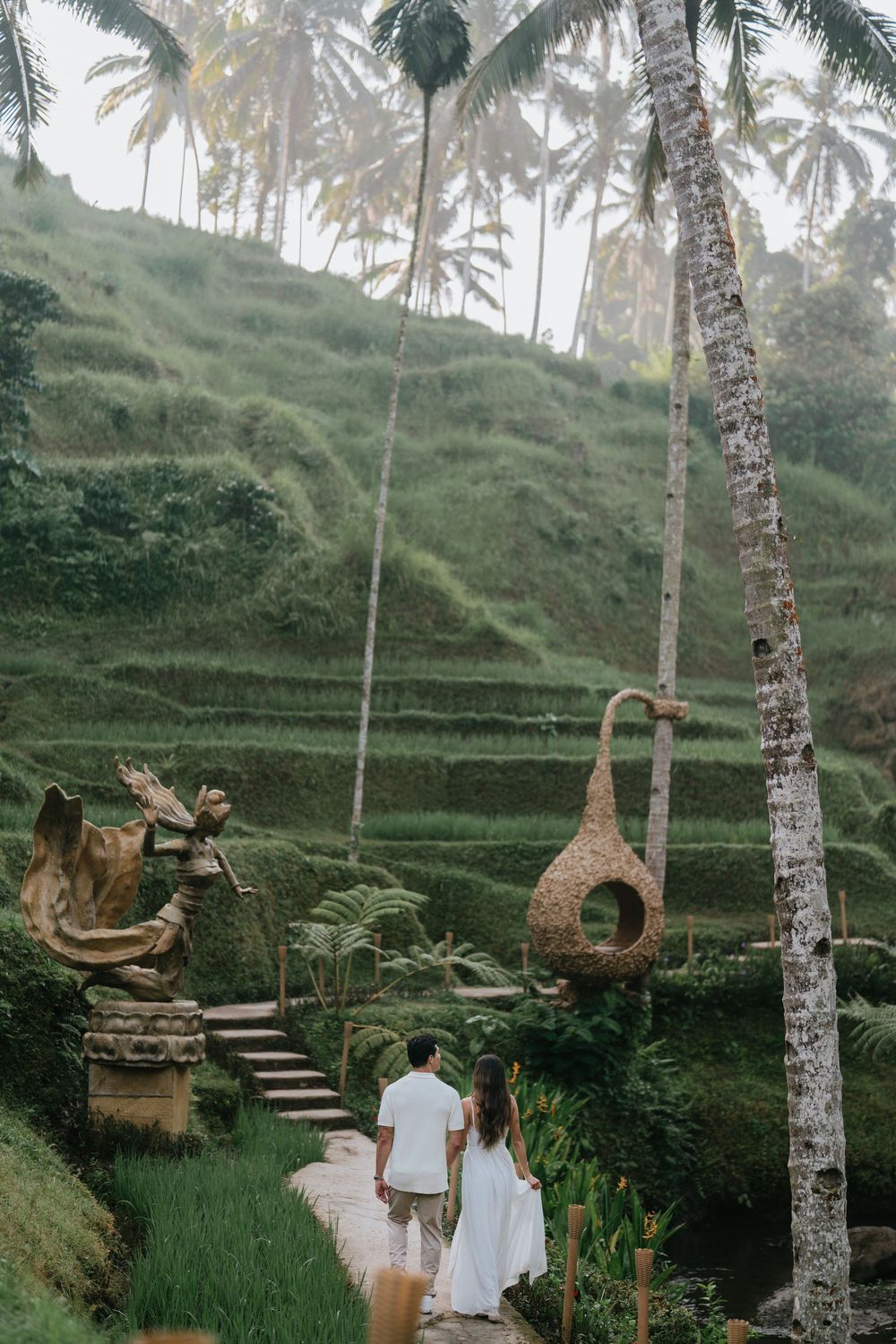 A couple walking down stone steps among rice terraces with decorative sculptures in Bali.