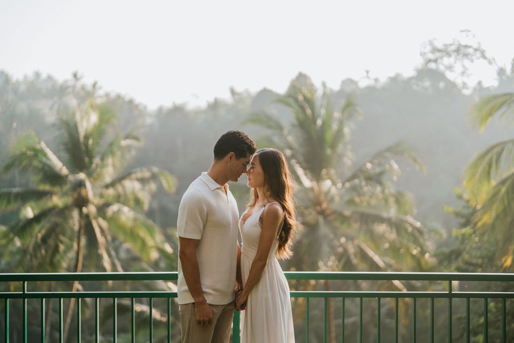 Couple in white outfits share intimate moment on tropical balcony with palm trees.