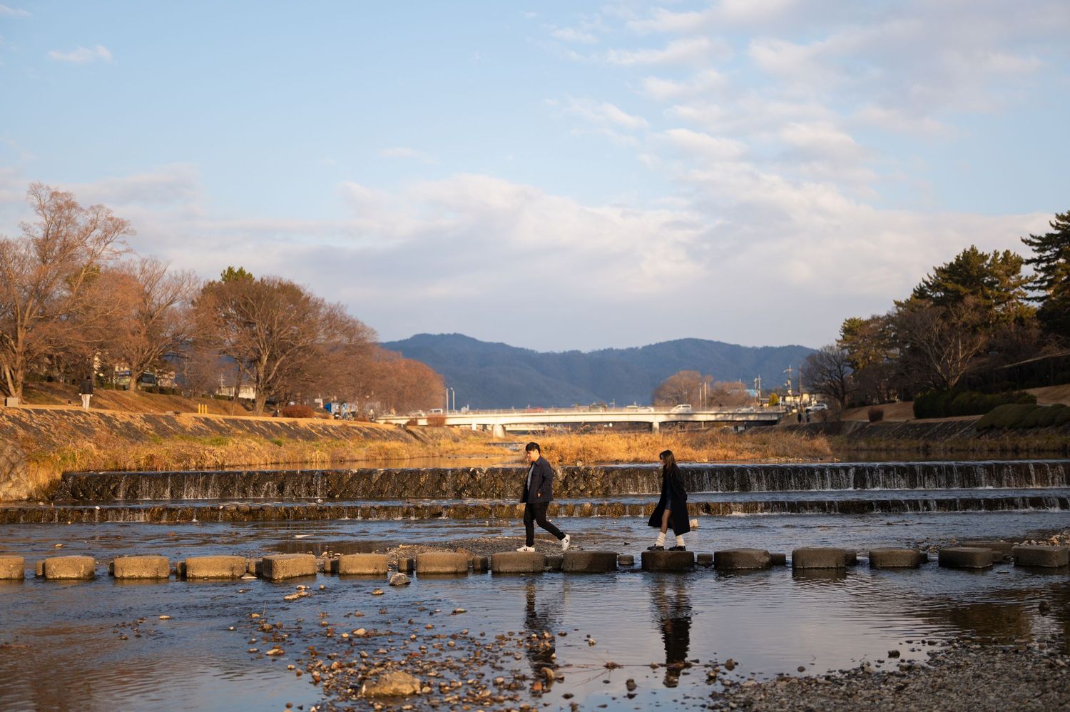 Couple crossing stepping stones over a shallow river with mountains in the background in Japan.