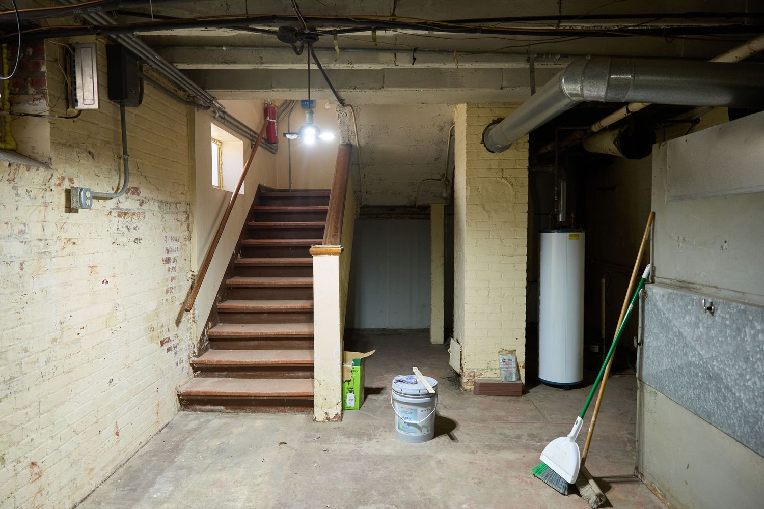 Unfinished basement space with exposed pipes, concrete walls, and wooden staircase.