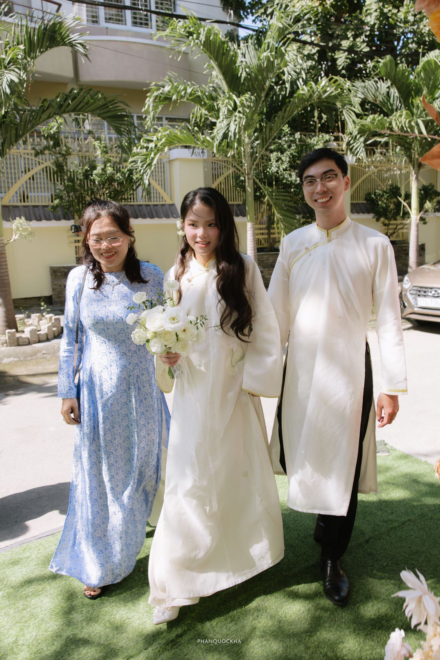 Vietnamese bride in white ao dai poses with bouquet between two family members outdoors.