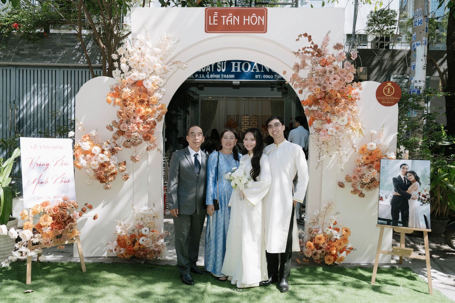 Couple in white ao dai posing at decorated arch entrance of Vietnamese wedding ceremony venue.