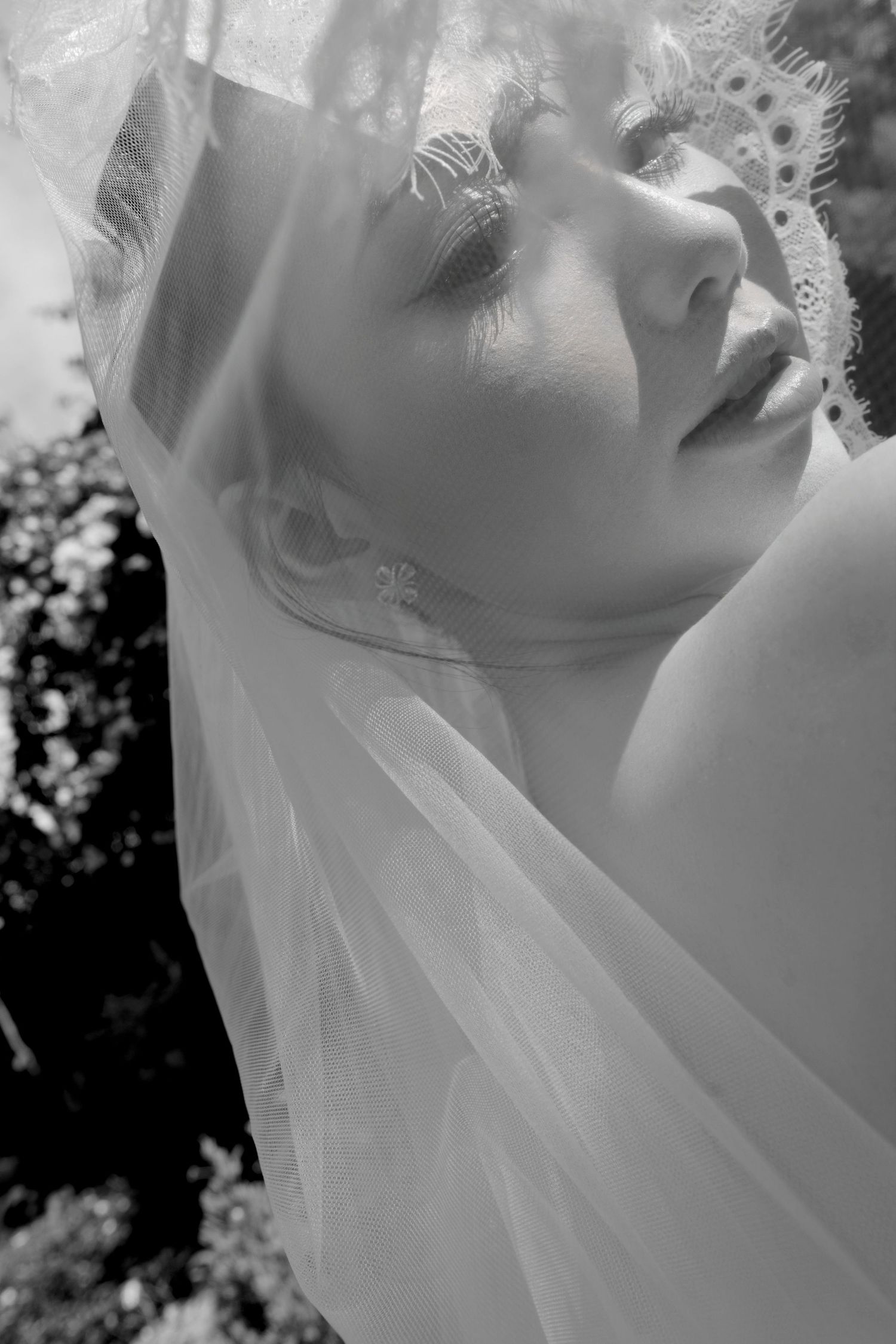 Close-up black and white portrait of a bride with a lace veil draped over her face, looking upward.