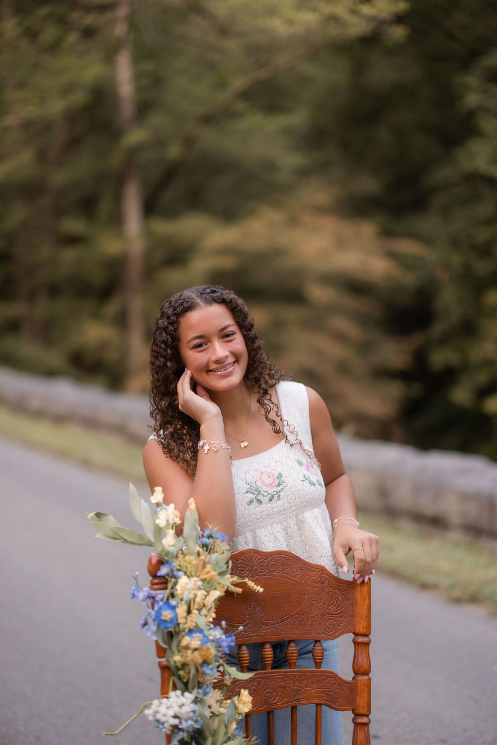 Someone in a white lace wedding dress sits on a wooden chair outdoors while holding a colorful bouquet and laughing joyfully.