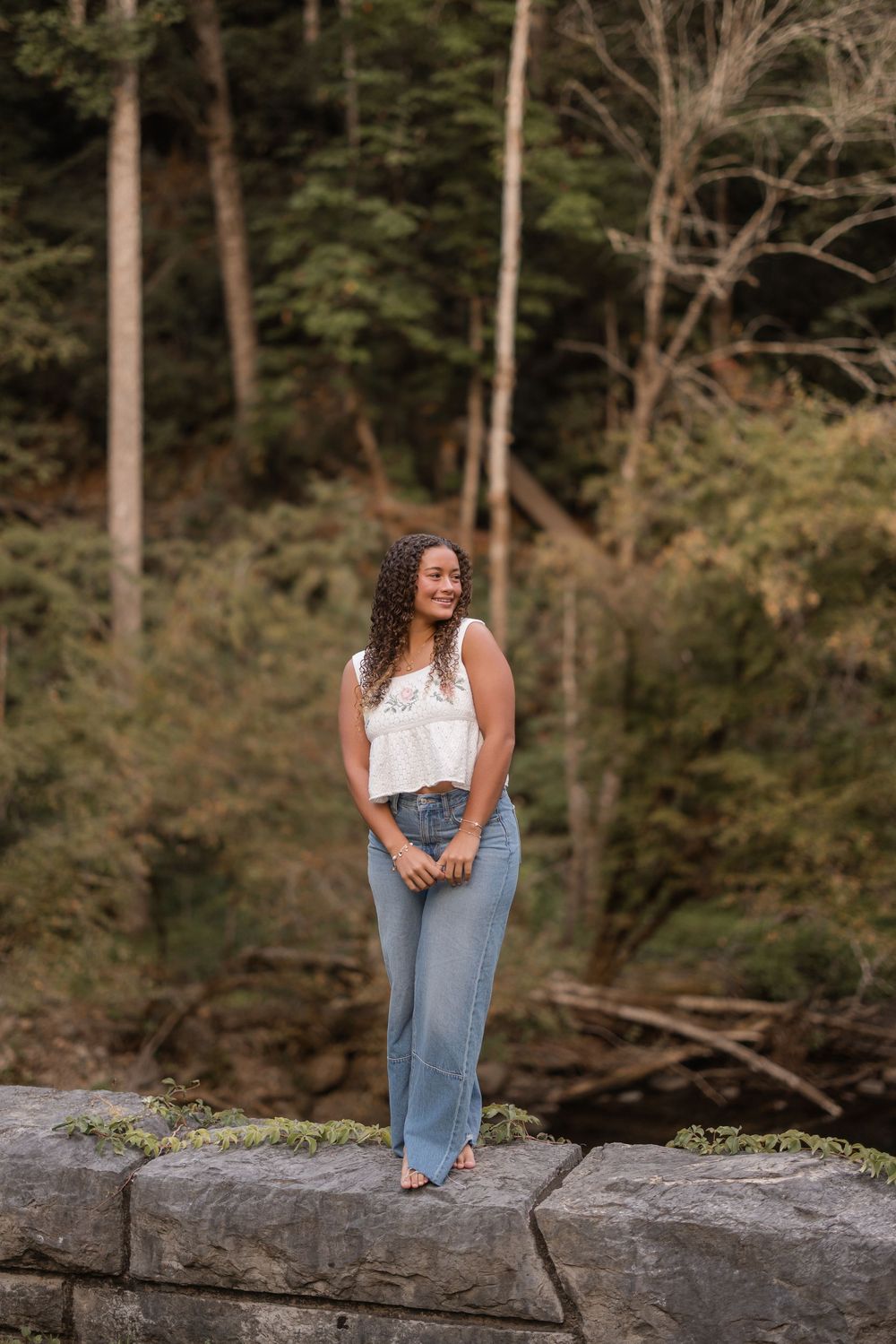 A dreamy outdoor portrait with someone sitting casually on a large rock in jeans and a white top against a wooded forest.