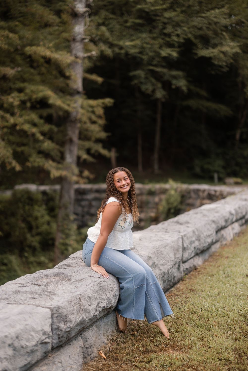 Someone in casual attire sits along a rustic stone wall with trees and greenery in the background.