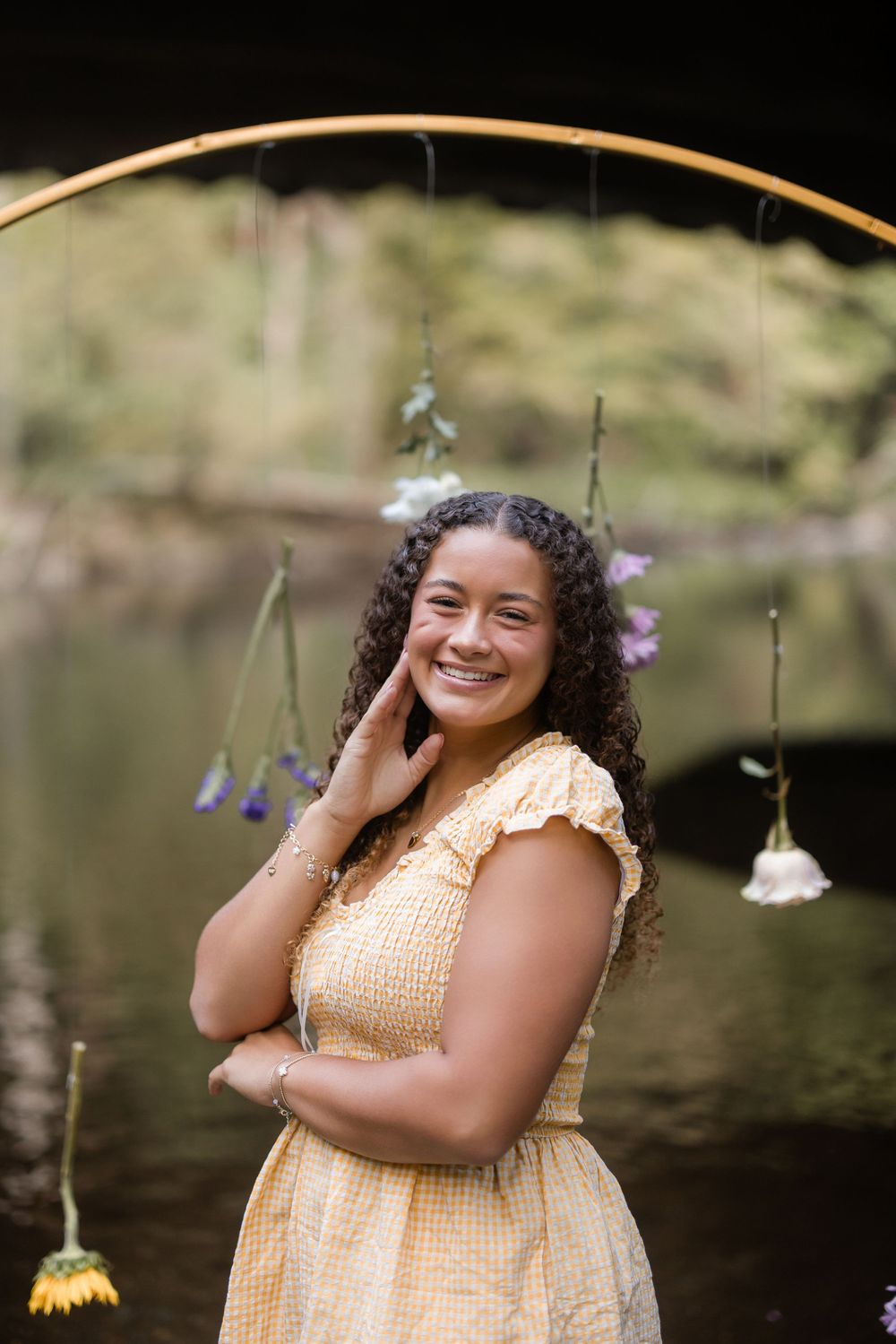A figure in a yellow sleeveless dress poses outdoors by a pond with hanging floral decorations.