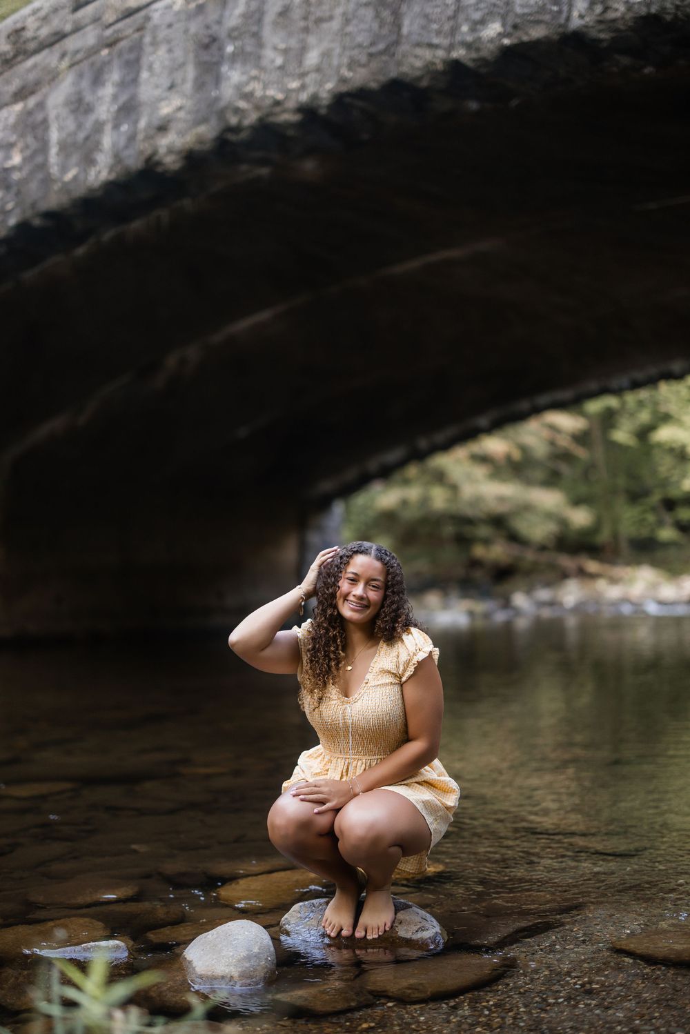 A person in a yellow dress crouches by the water's edge under a rocky overhang in a natural outdoor setting.