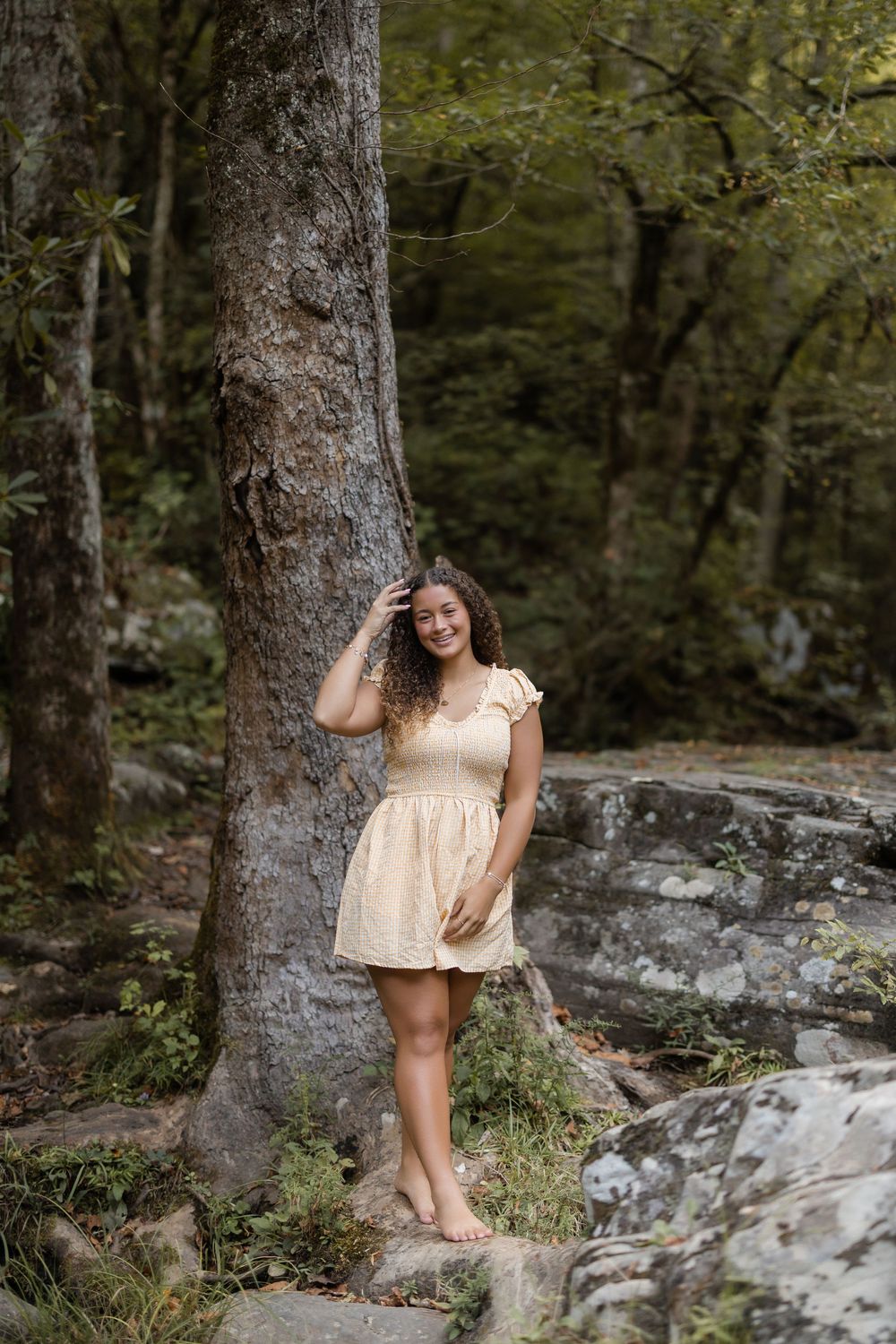 A person wearing a light-colored dress poses near large tree trunks and rocks in a serene forest setting.