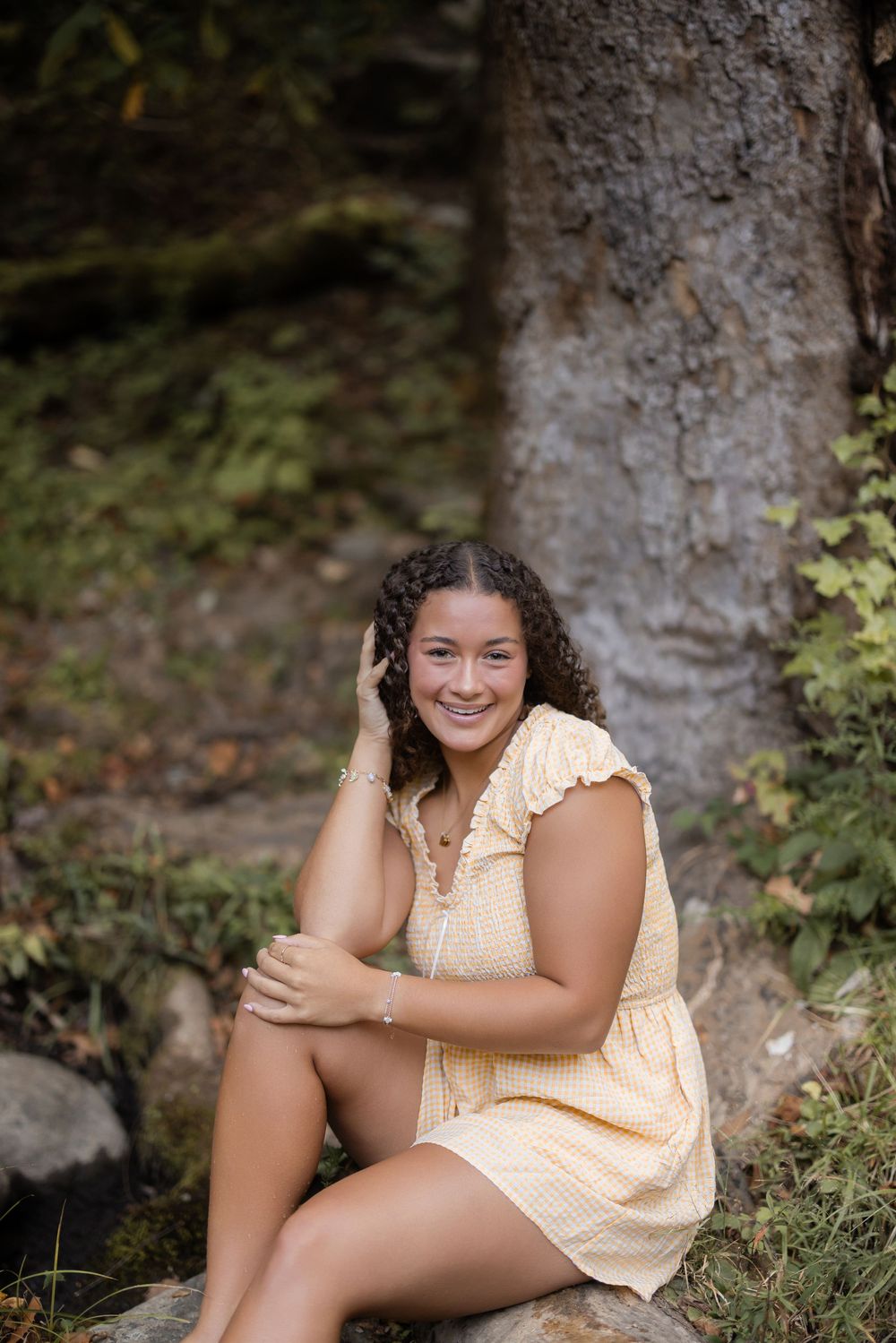 Young person in peach-colored dress sits outdoors against tree trunk background in natural lighting.