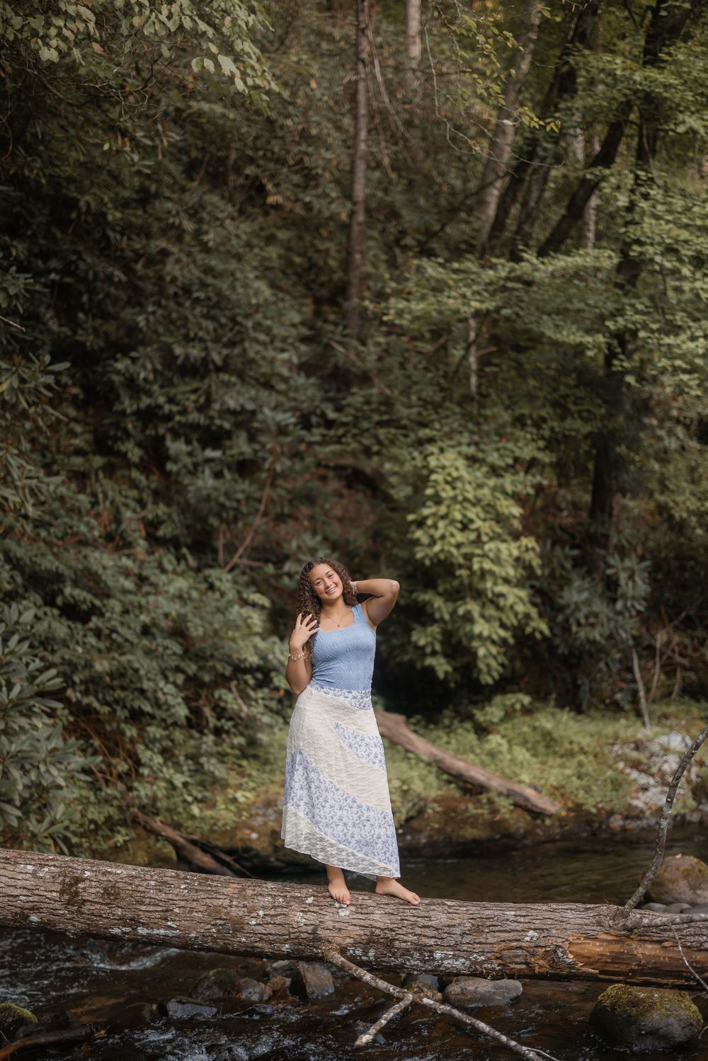A person in a white floral dress perches gracefully on a fallen log over a peaceful forest stream on a summer day.