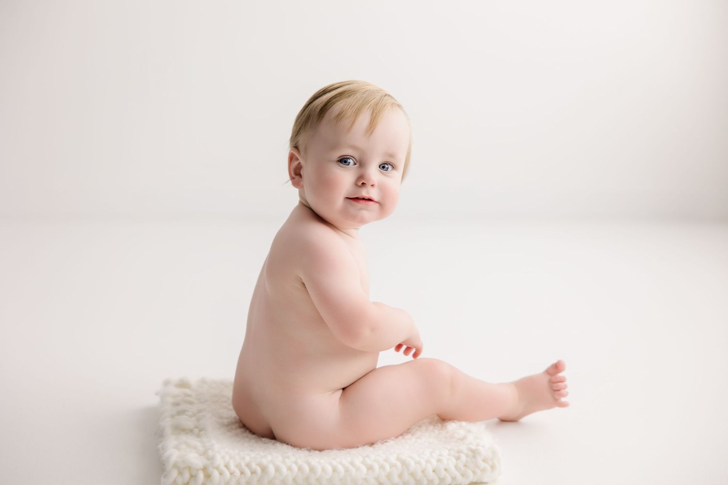 Baby wearing white outfit sits on white backdrop during minimalist photo session.