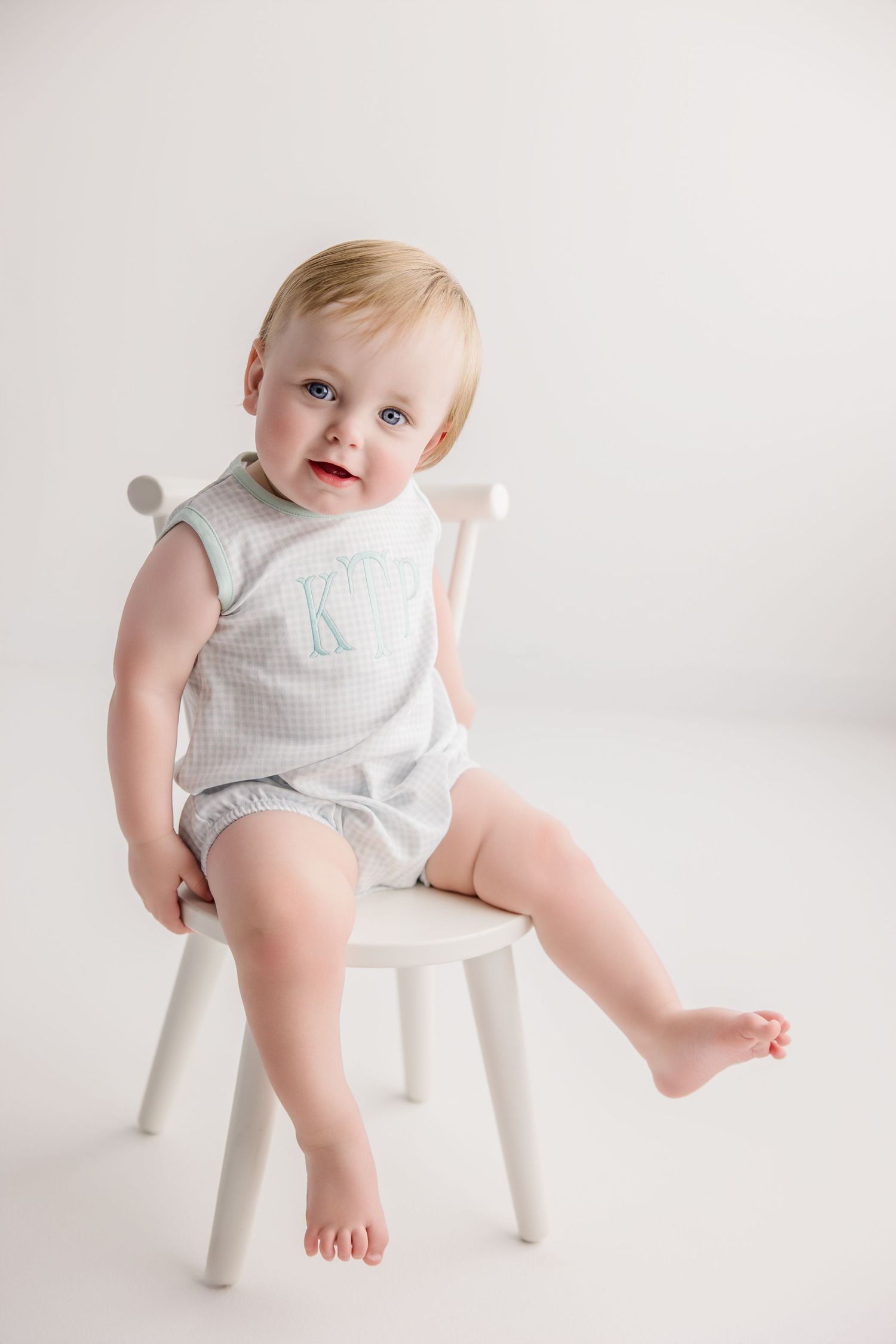 A baby in a light colored romper sits playfully on a small white wooden stool against a white background.