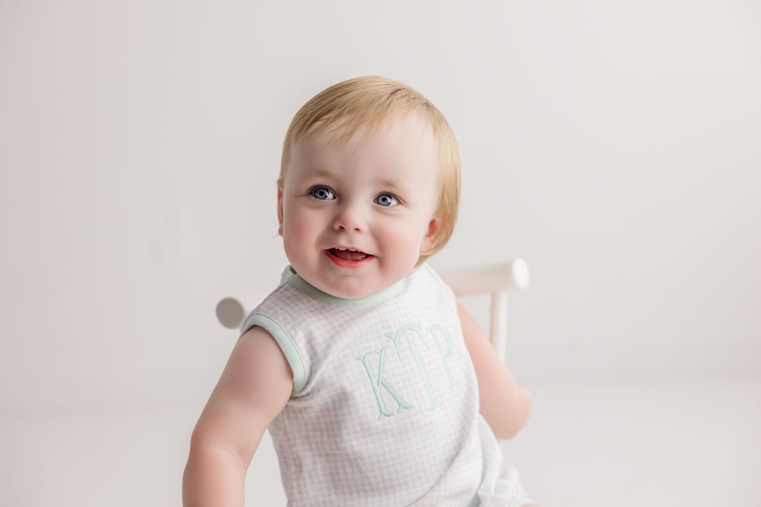 A happy baby with blonde hair wearing a white outfit sits against a light-colored background.