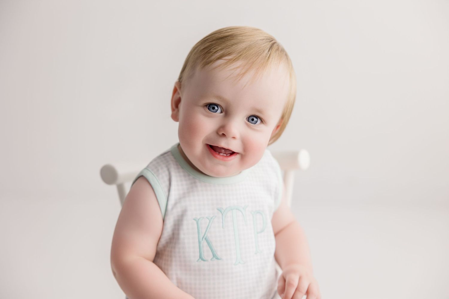 Baby in soft white outfit smiles while sitting against white background.