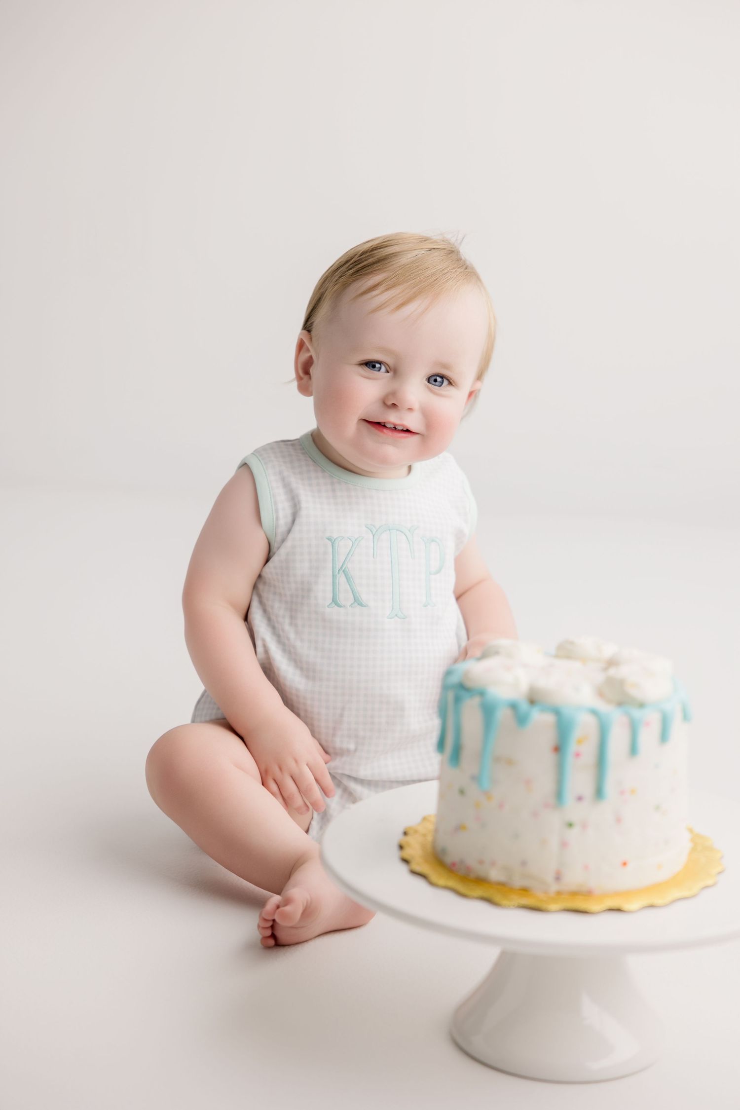 White cake with blue decorations sits on a cake stand while an infant in a white outfit looks on curiously.