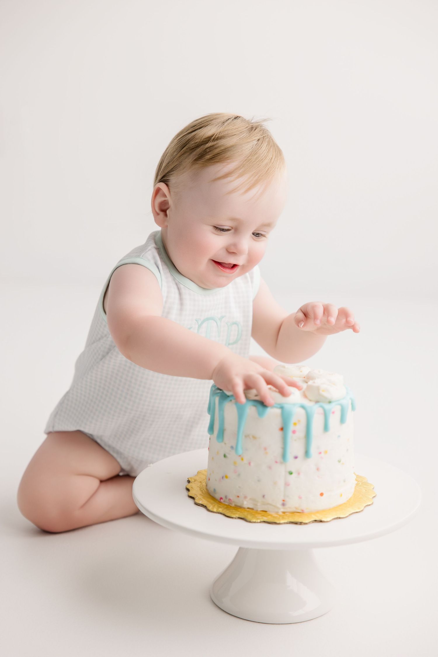 Baby in white sleeveless outfit reaching for white birthday cake with blue drips.