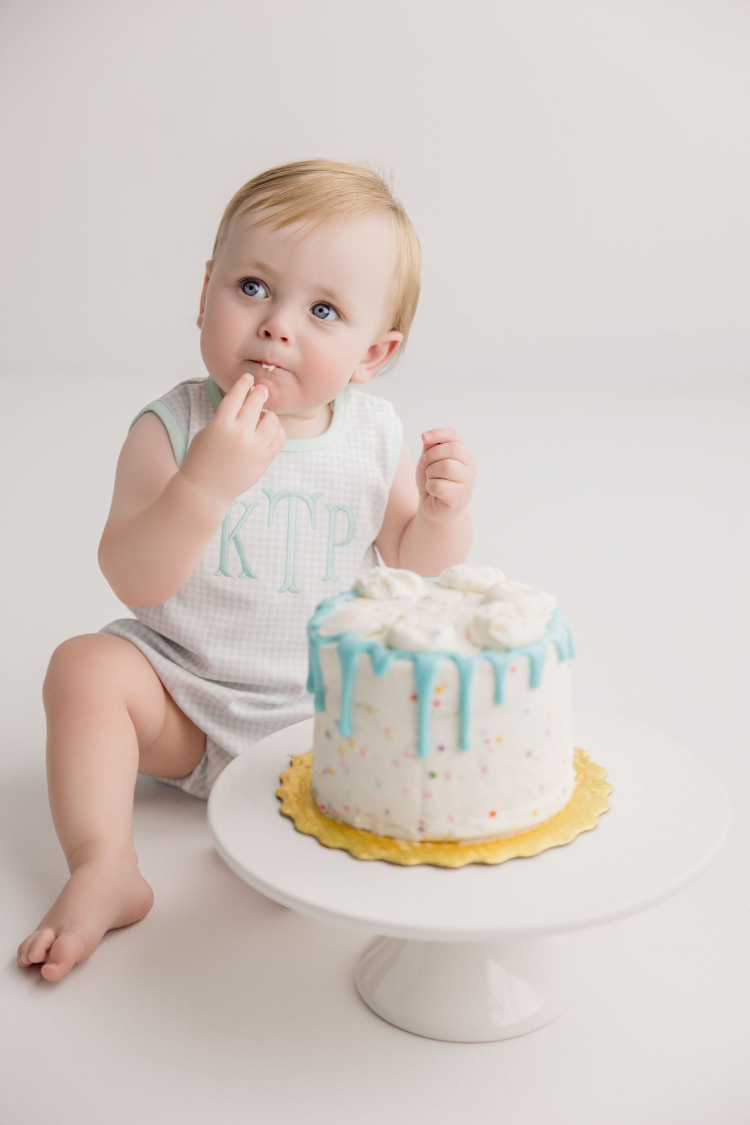 Four sequential photos showing a baby in white reaching for and tasting their first birthday cake on a white cake stand.