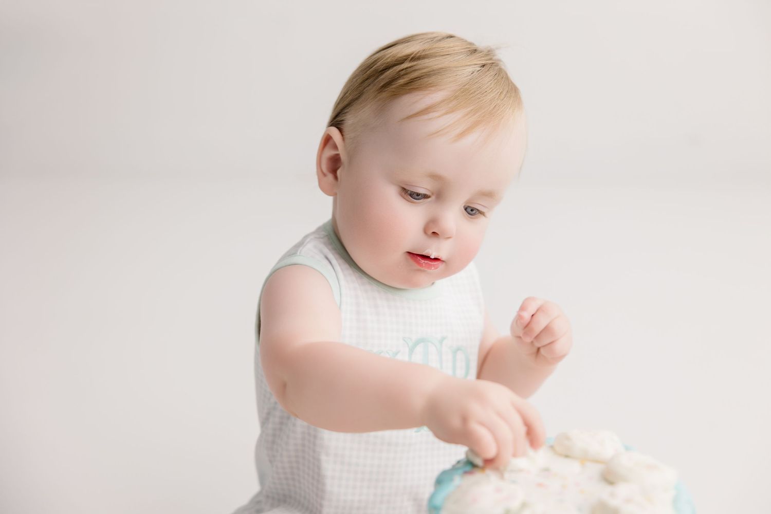 Baby in white dress touches cake during cake smash photo session against white background.