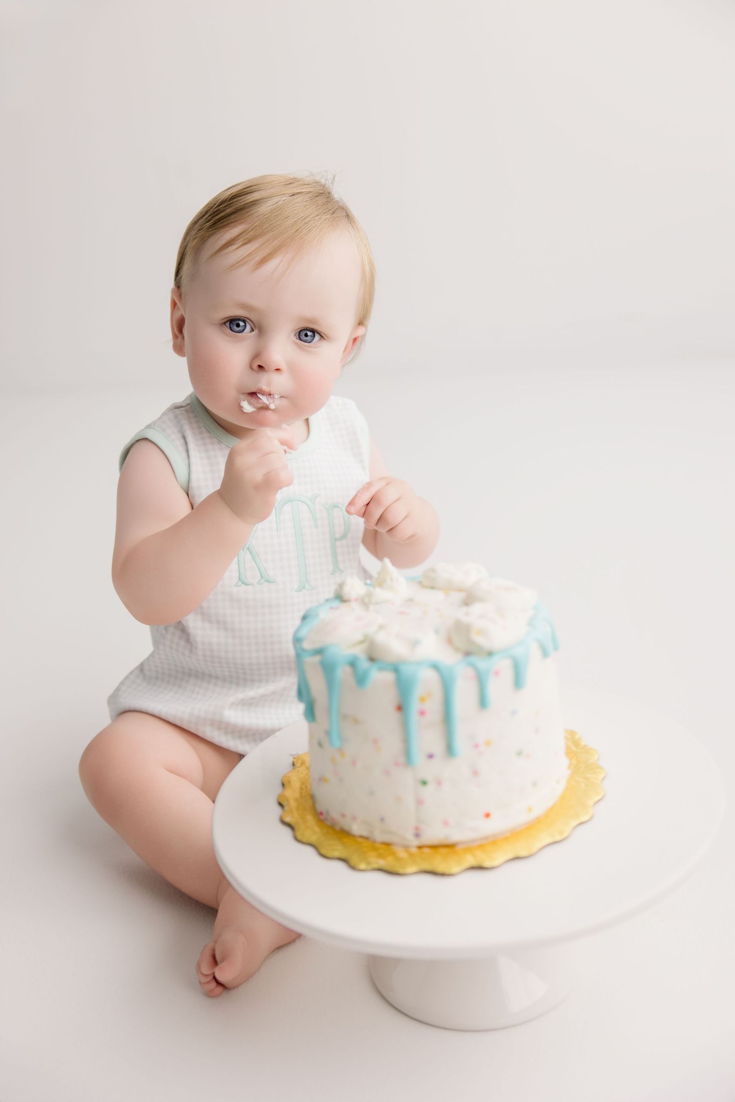A first birthday cake smash photo series showing a baby with a white and blue decorated cake on a cake stand.