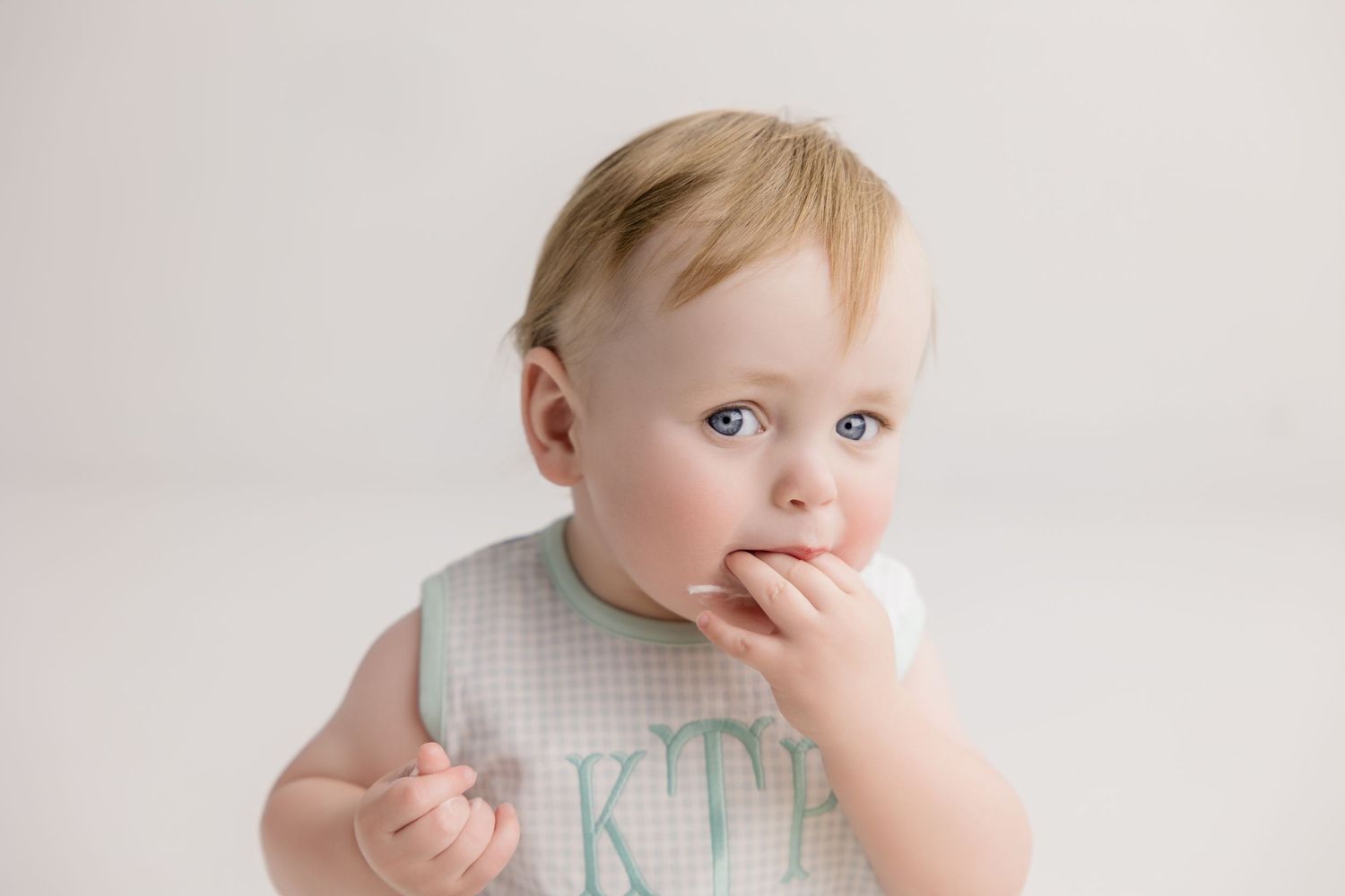 Baby in light colored outfit looking thoughtfully while touching their face against neutral background.