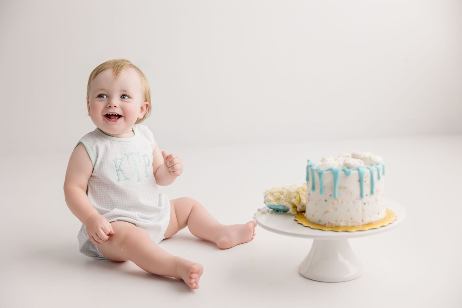 Baby wearing white sits in against white backdrop near blue and white cake during cake smash photo session.