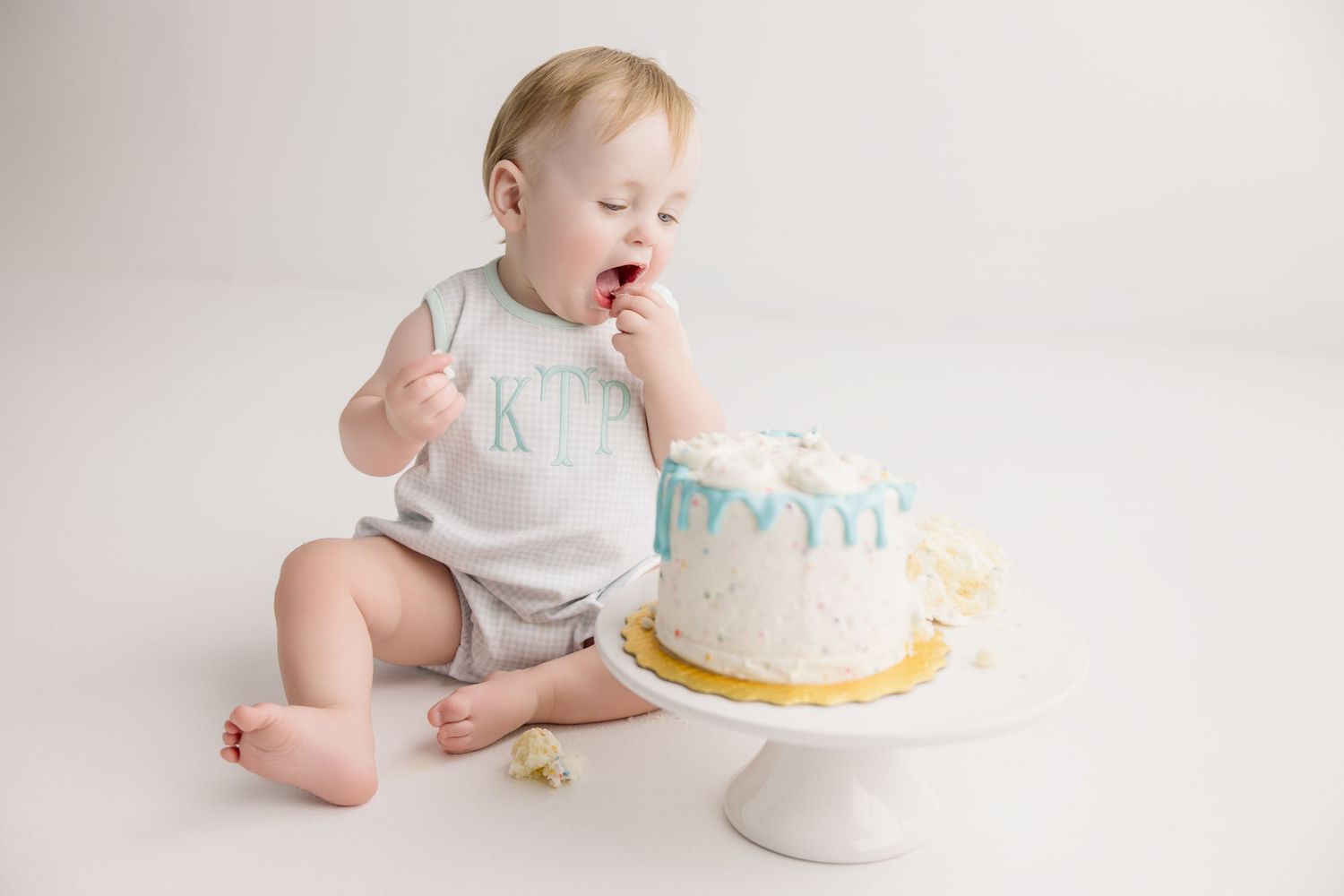 Baby reaches for small white cake during first birthday cake smash photography session.
