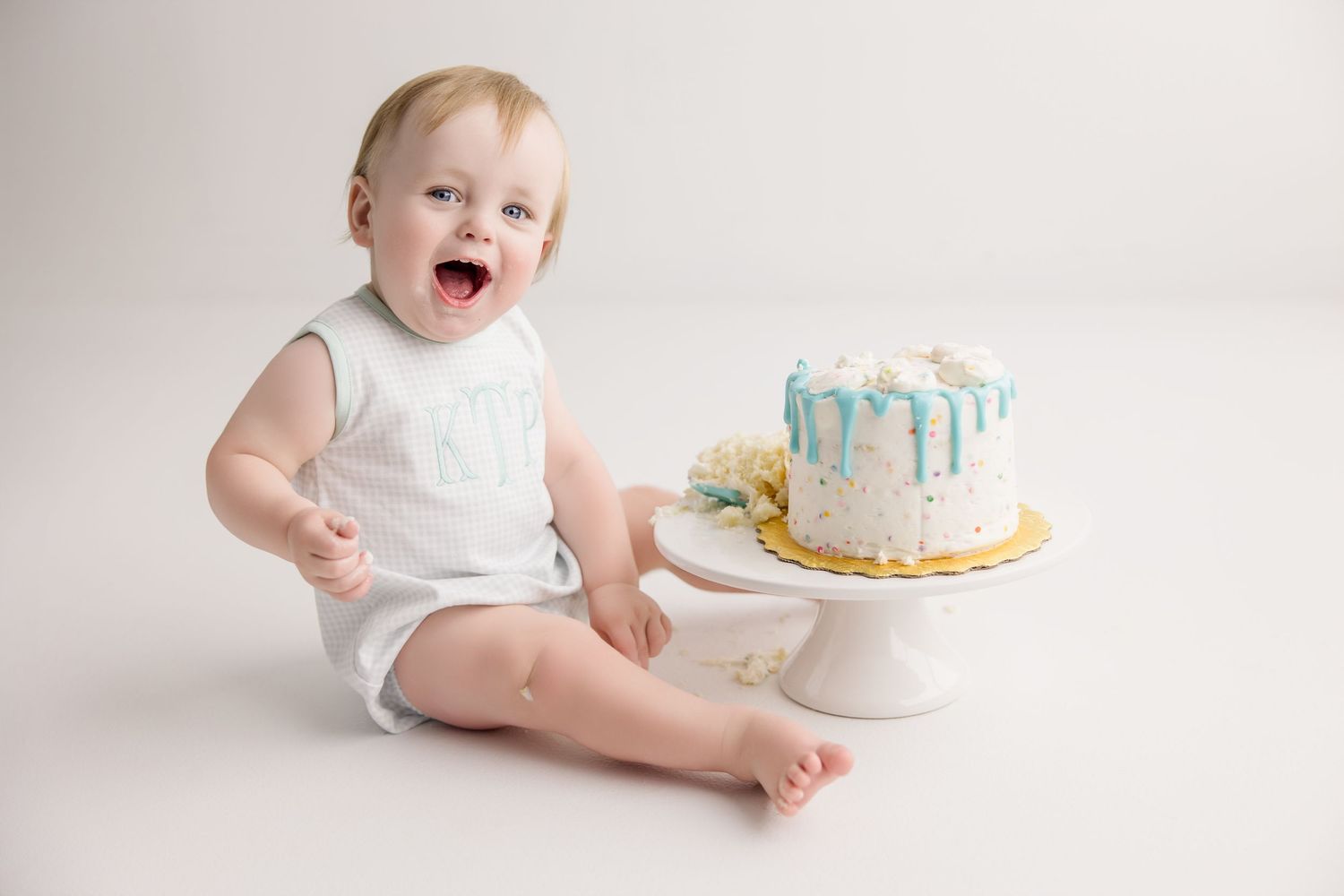 Baby leaning towards white cake with blue drips on pedestal during cake smash session.