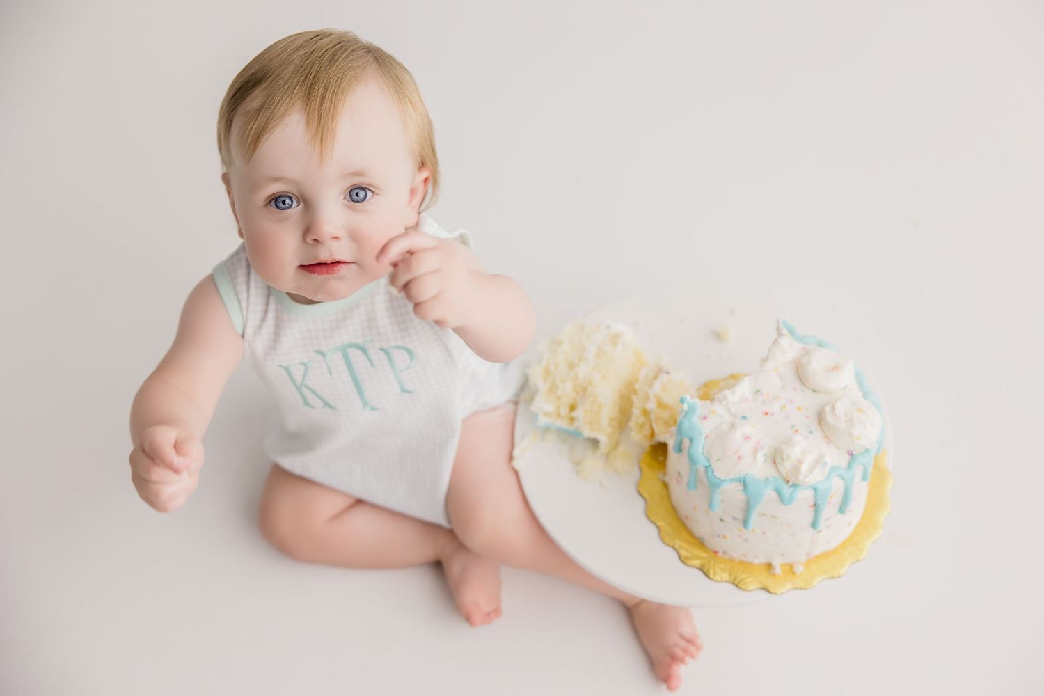 Baby in white outfit sitting next to decorated cake with blue and peach frosting.