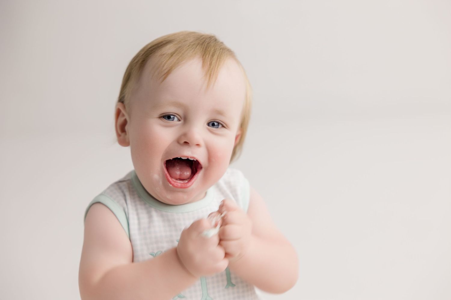 Joyful baby wearing light colored outfit laughing and clapping against white background.