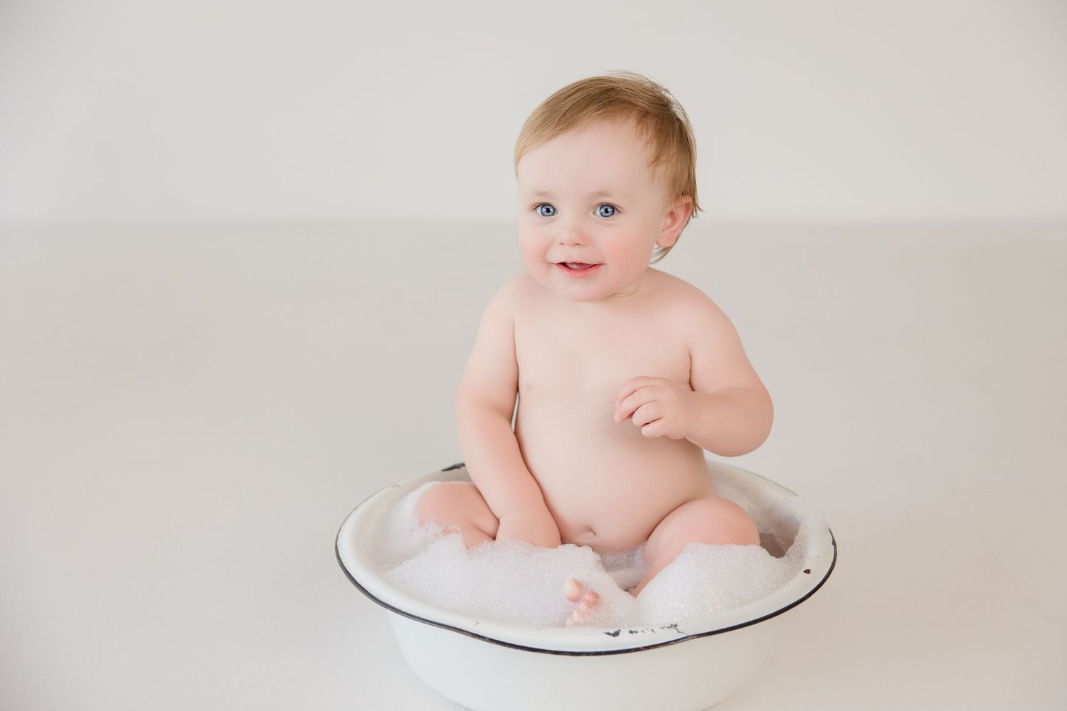 Happy baby sits in white bowl during minimalist portrait session.
