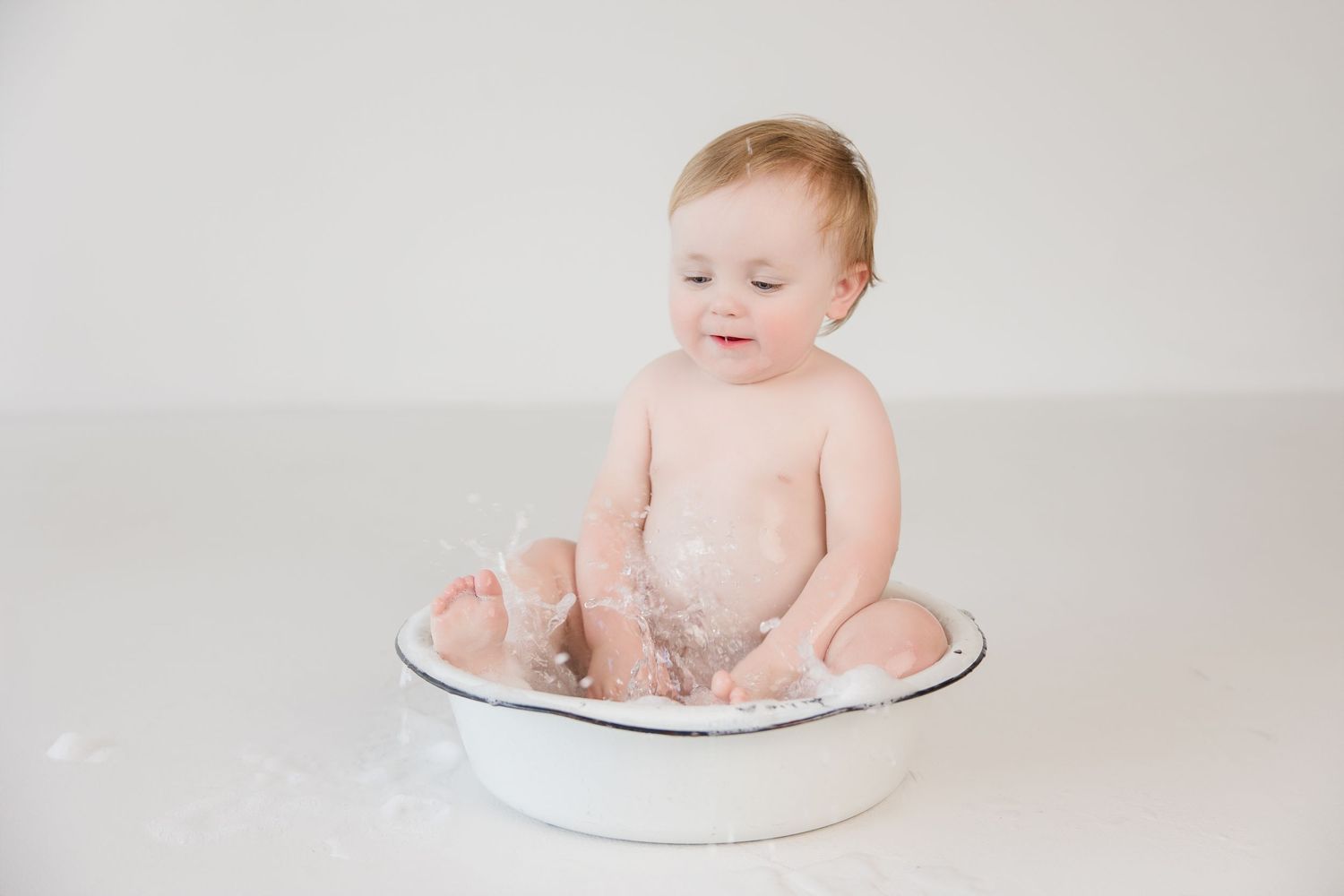 An infant sits in a white basin for bath time while splashing water during a fun photo session.